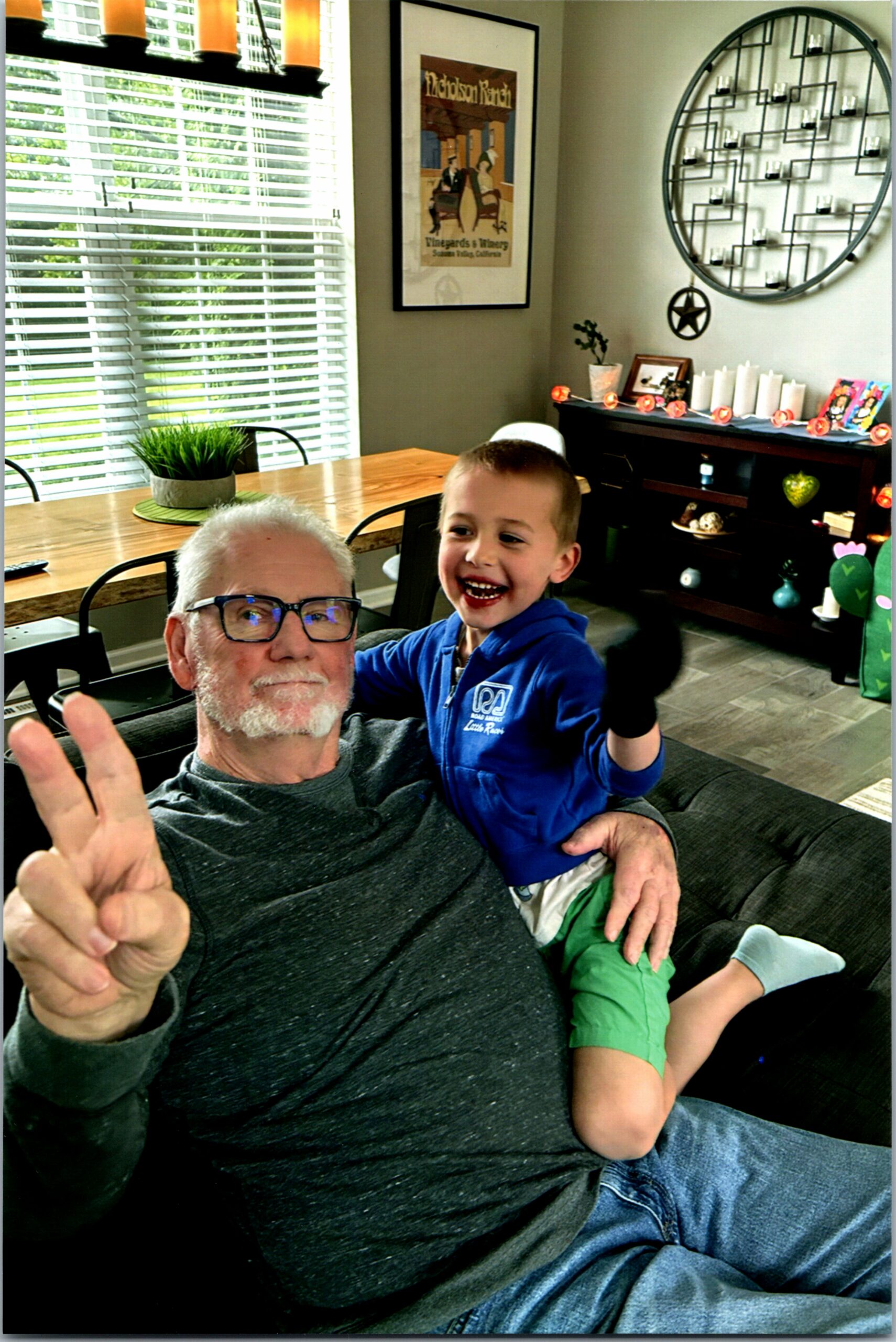 Grandfather and young boy smiling on a sofa in a bright living room, both waving at the camera.