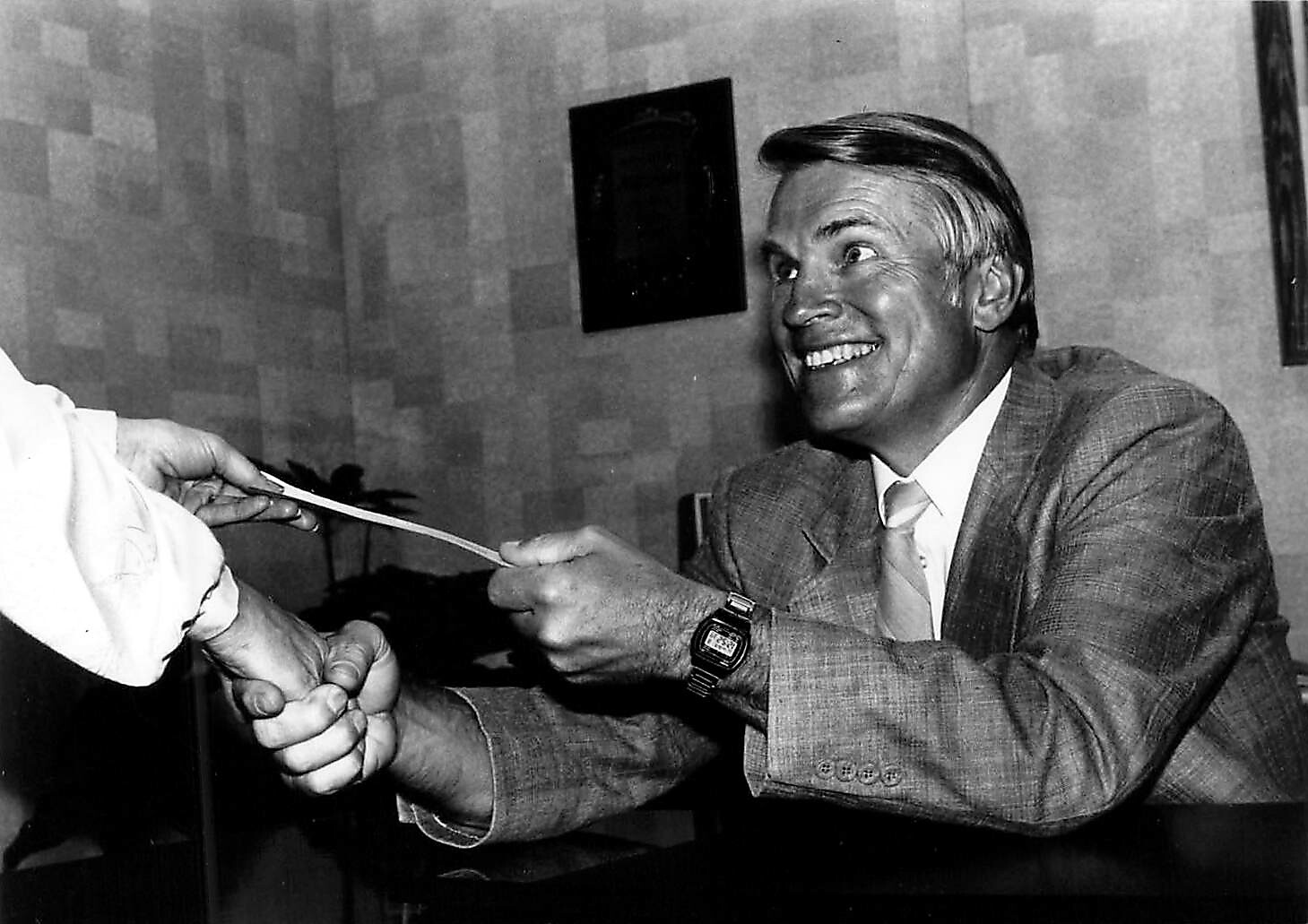 Smiling businessman in a suit hands a document to another person across a desk.