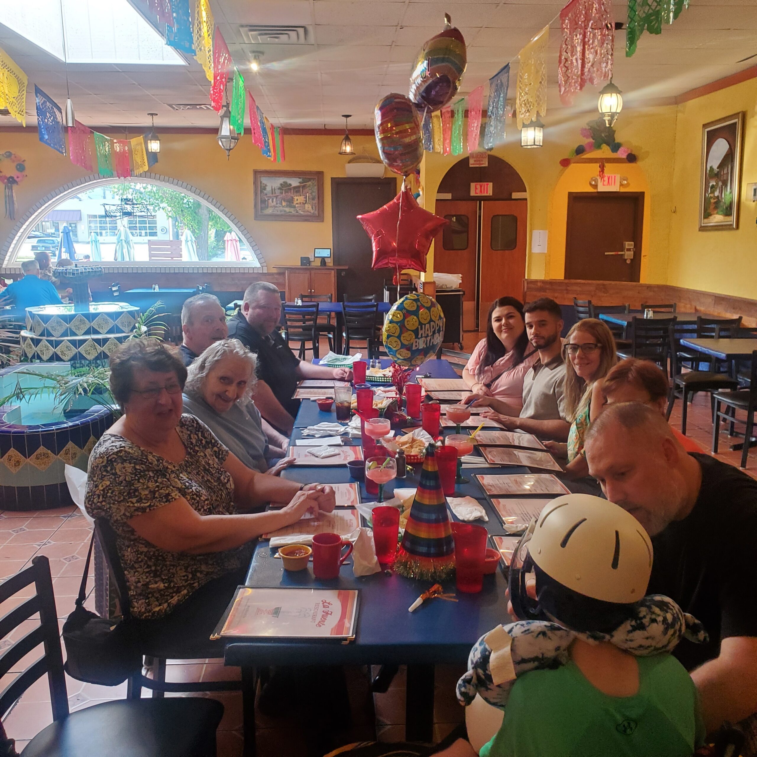 People seated at a long table in a festively decorated restaurant with birthday balloons overhead.