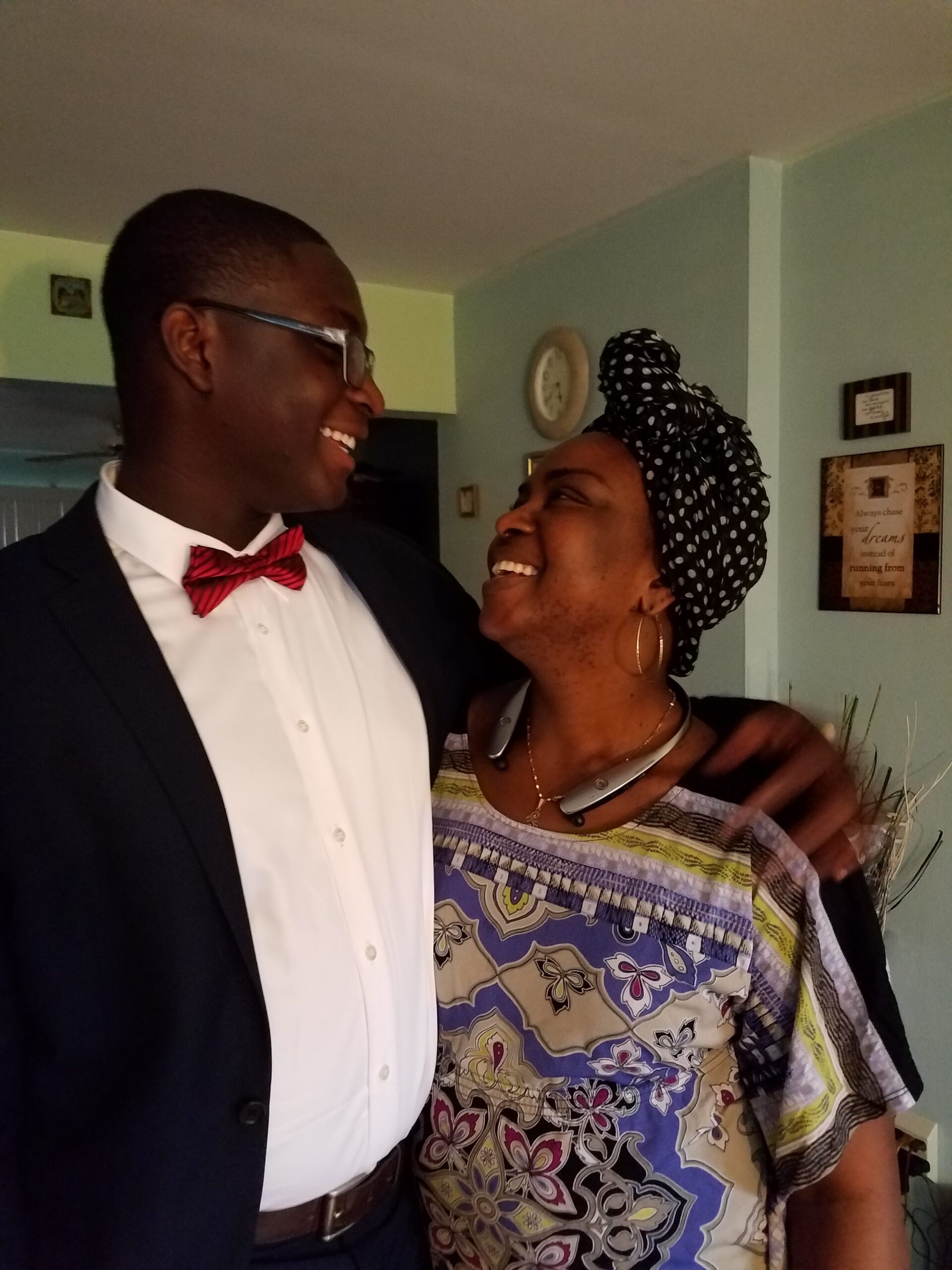 Two people smile at each other indoors: a man in a suit with a red bow tie and a woman wearing a patterned dress and a black-and-white headwrap.