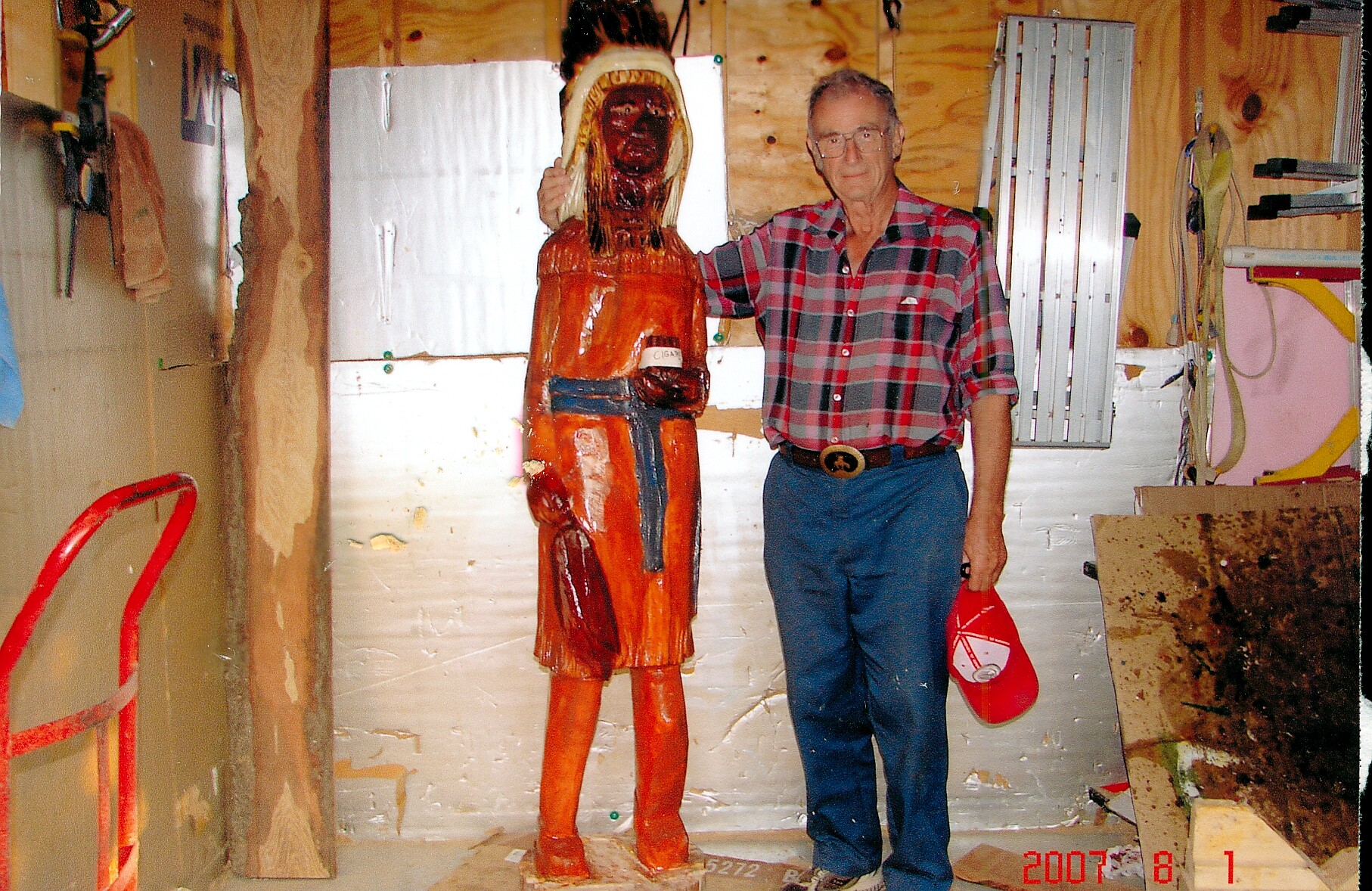 Older man in a red-and-blue plaid shirt stands beside a tall orange-painted wooden statue in a cluttered workshop.
