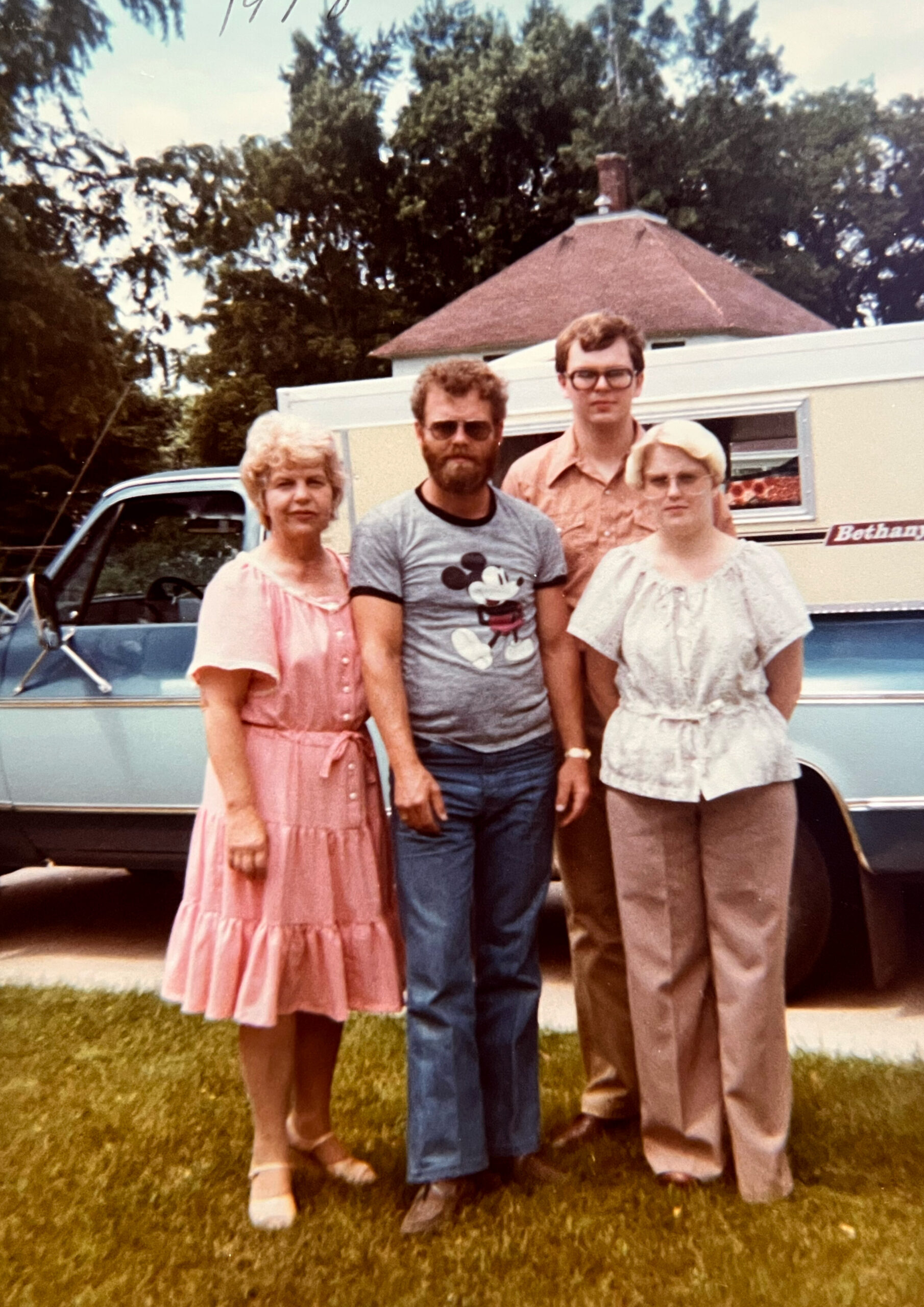 Four family members pose in front of a light blue vintage car and beige camper in a yard, likely from the 1970s or 1980s.