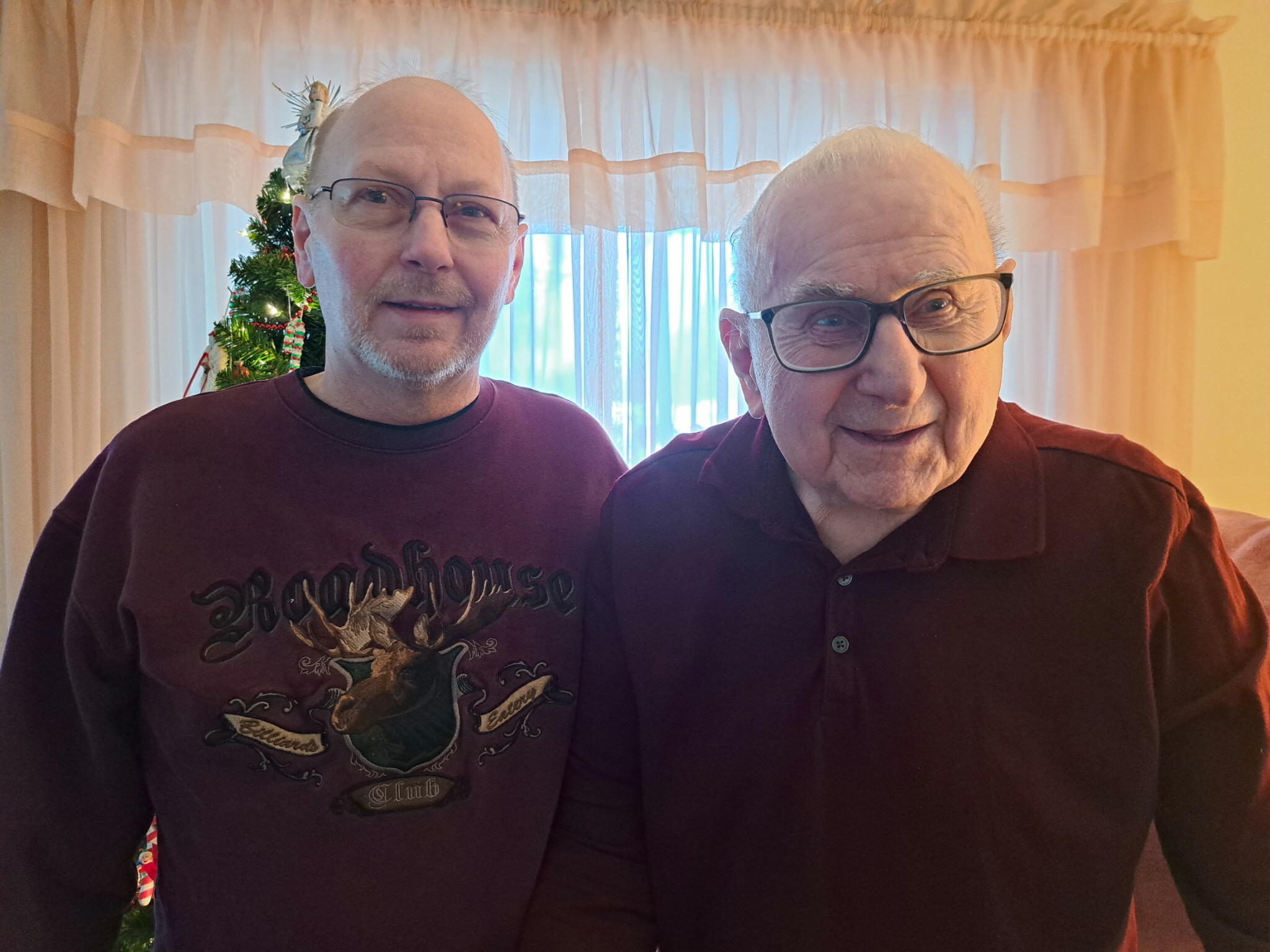 Two men pose for a selfie in a cozy living room with a decorated Christmas tree in the background, both wearing maroon shirts.