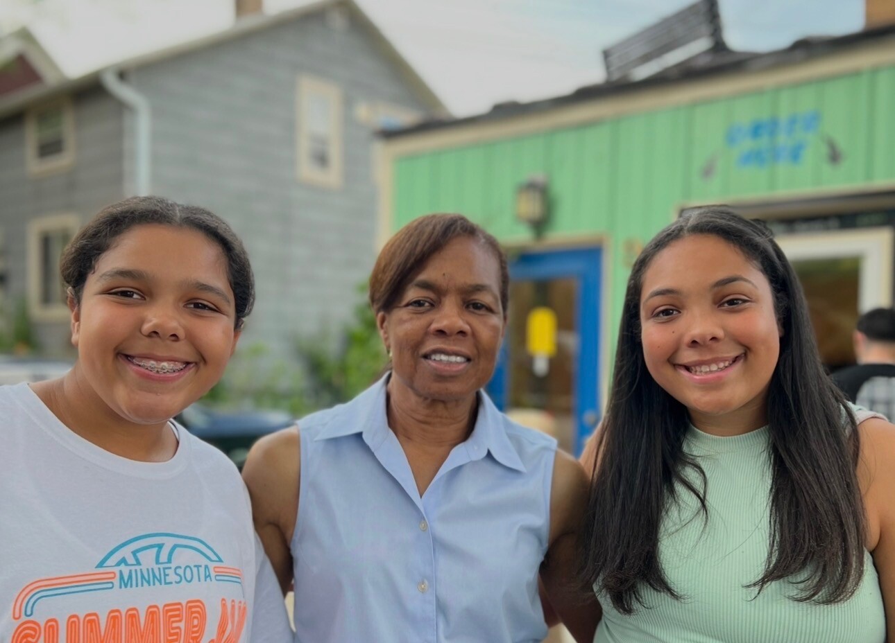 Three people smile for a group photo outdoors in front of colorful buildings, including a girl with braces on the left, an older woman in the middle, and a girl in a mint green top on the right.
