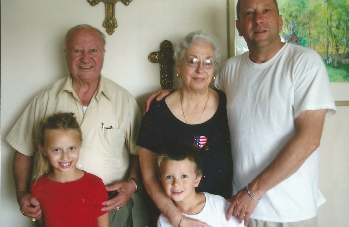 A multi-generational family portrait indoors: two grandparents with a man and two smiling children posing together.