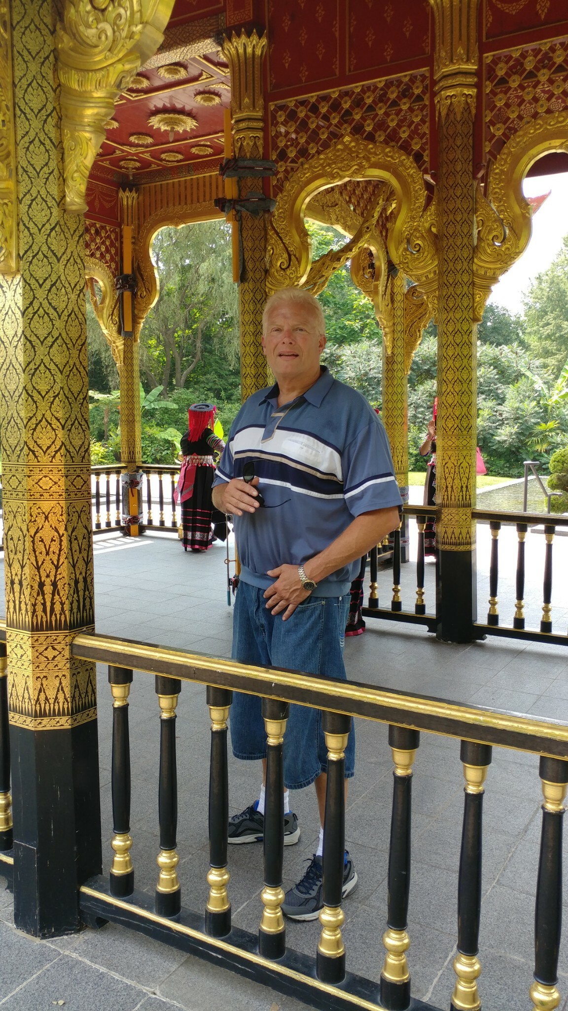 Older man in a blue striped polo and shorts stands inside a gold ornate pavilion with carved columns and railing.