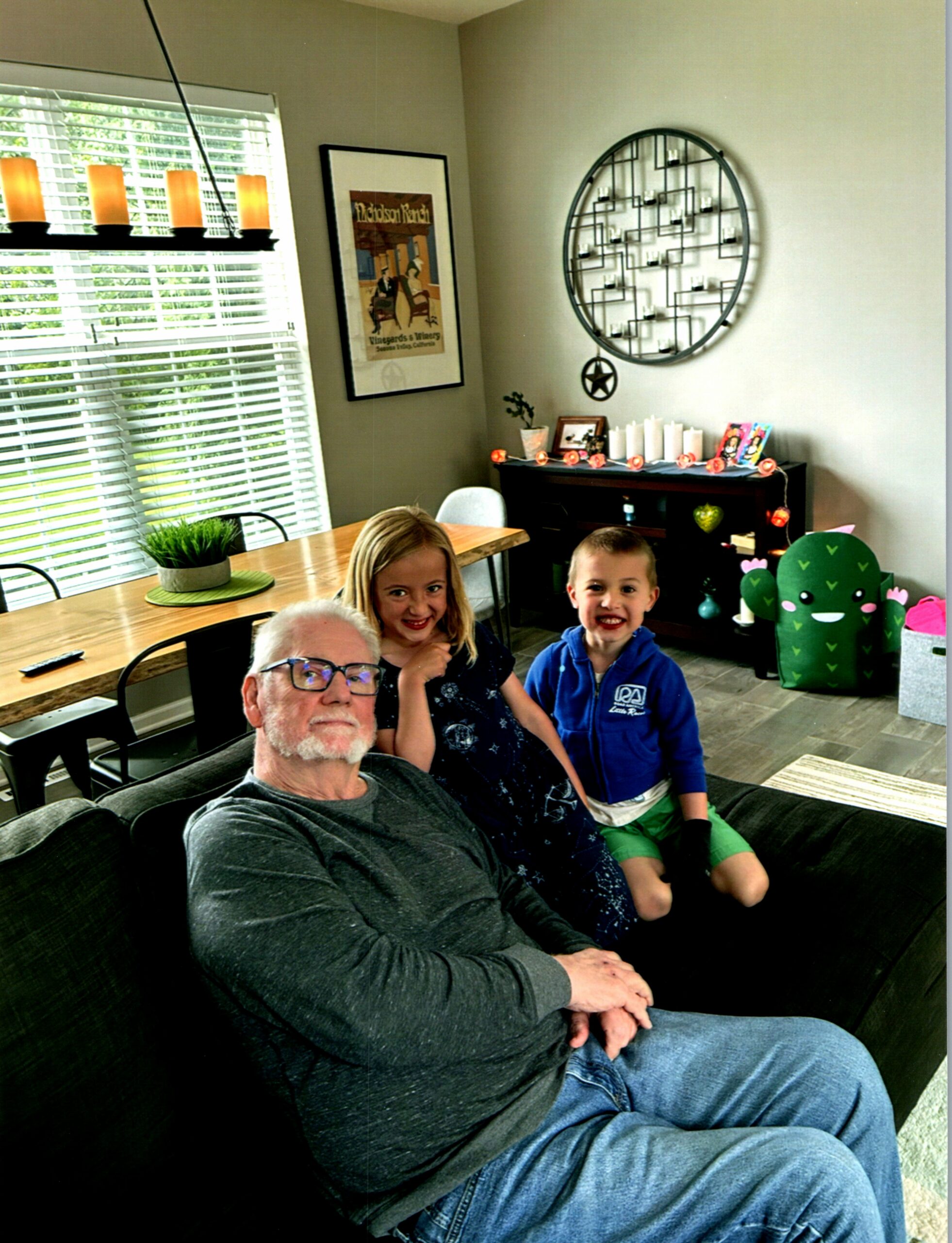 Family of four sitting on a dark sofa in a sunny living room, smiling at the camera with bright window blinds behind them.