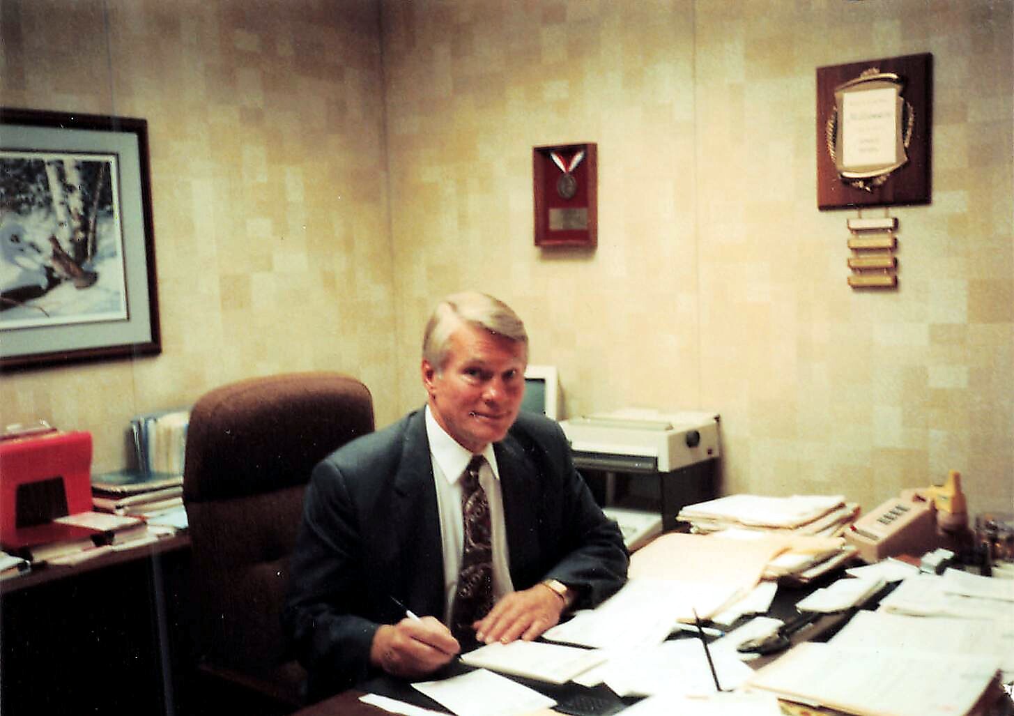 Man in a suit sits at a cluttered office desk, signing papers.