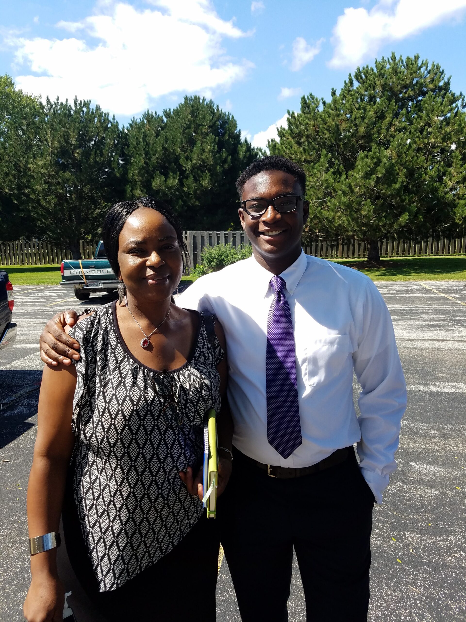Two people posing together outdoors, smiling with arms around each other in a parking lot, trees and blue sky behind them.