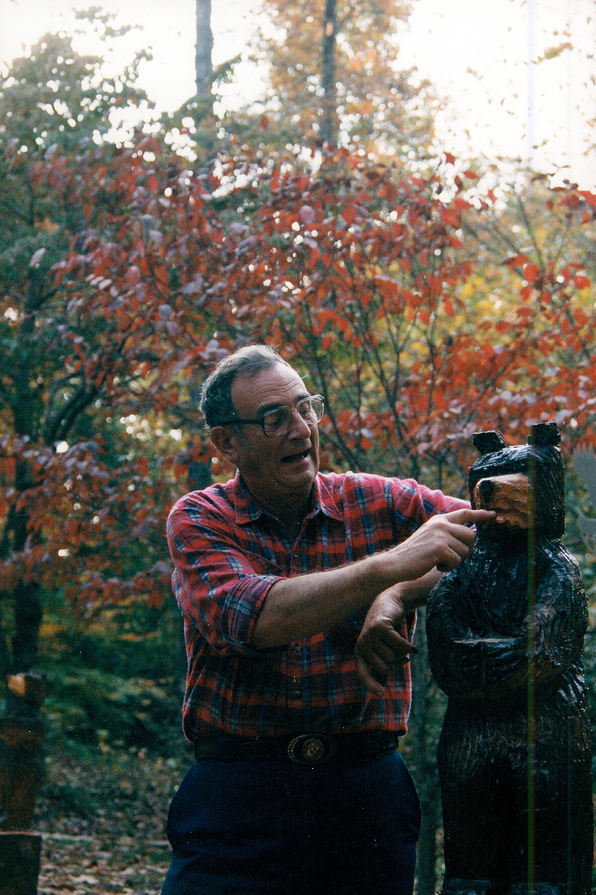 Older man wearing glasses and a red plaid shirt points at a carved wooden statue outdoors in a fall garden.