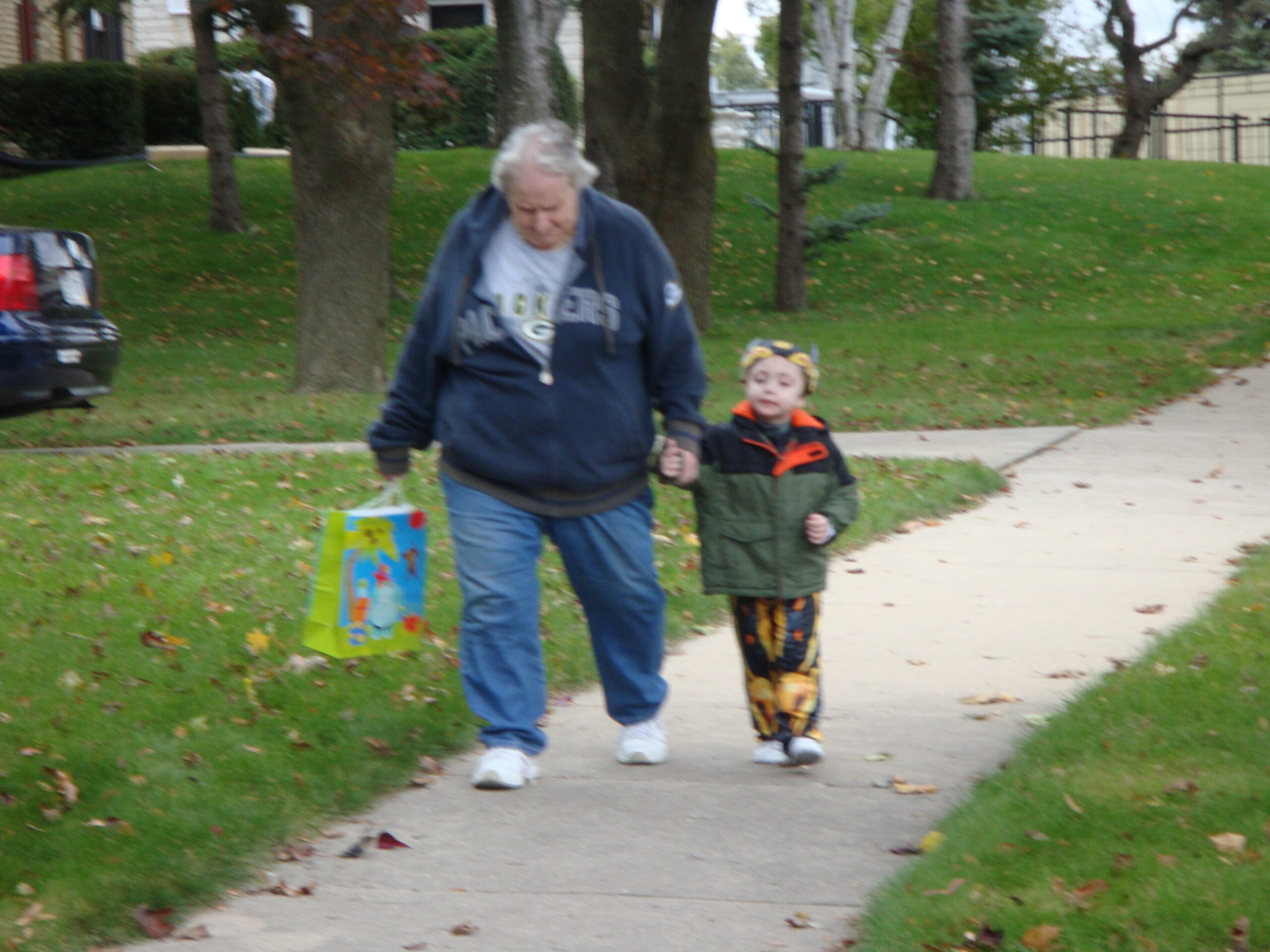 Older man in a blue jacket walking hand-in-hand with a young child on a sidewalk in a park-like neighborhood; autumn leaves on the ground.