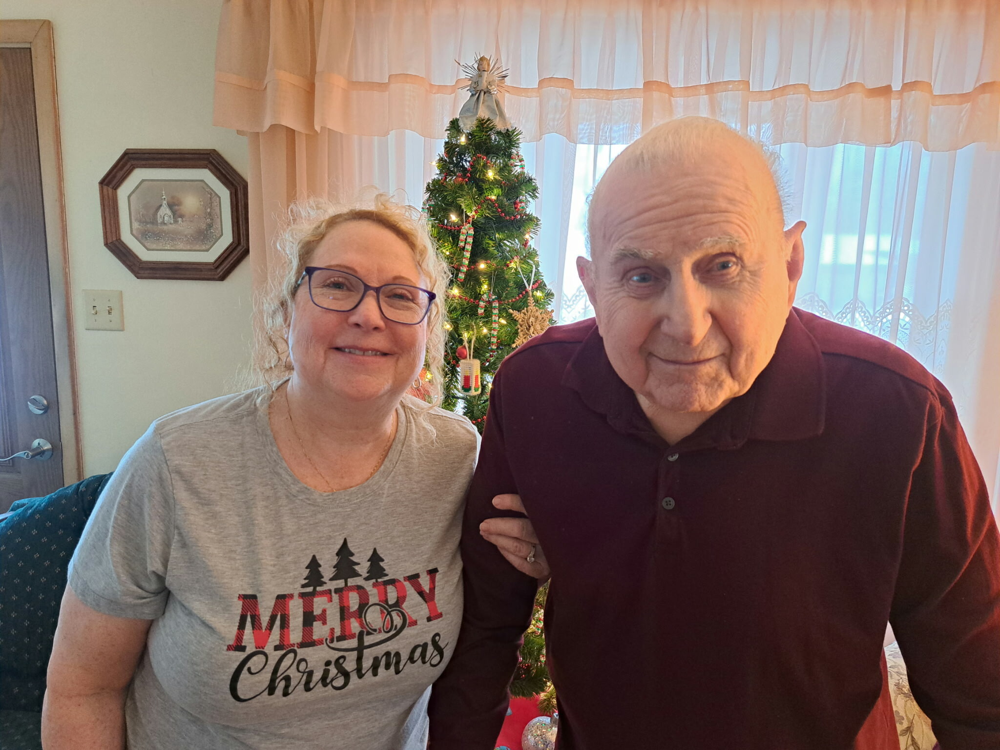 Smiling woman and man standing indoors with a decorated Christmas tree behind them.