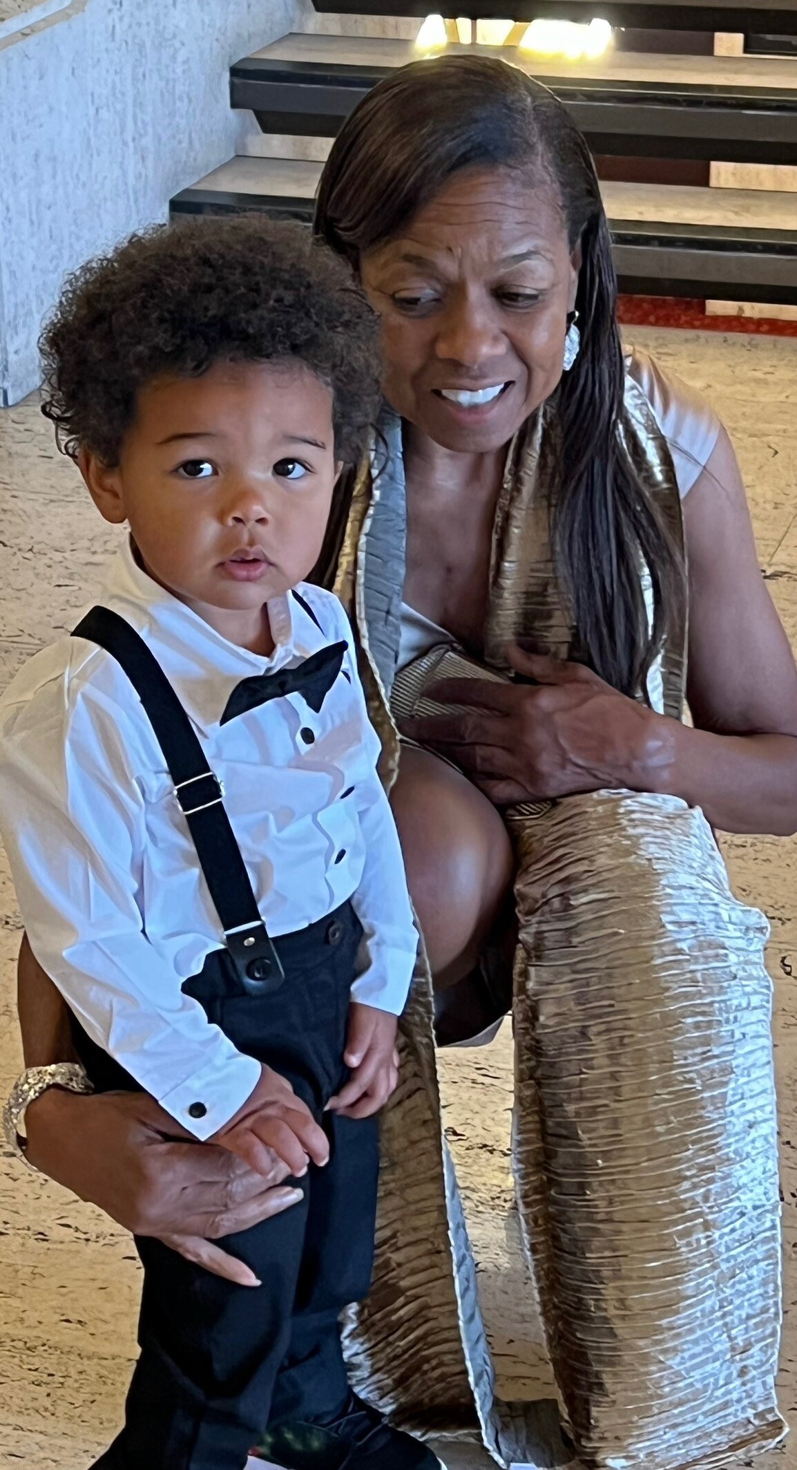 A smiling woman in a gold dress beside a toddler in a white shirt, black suspenders, and a bow tie, posed together indoors.