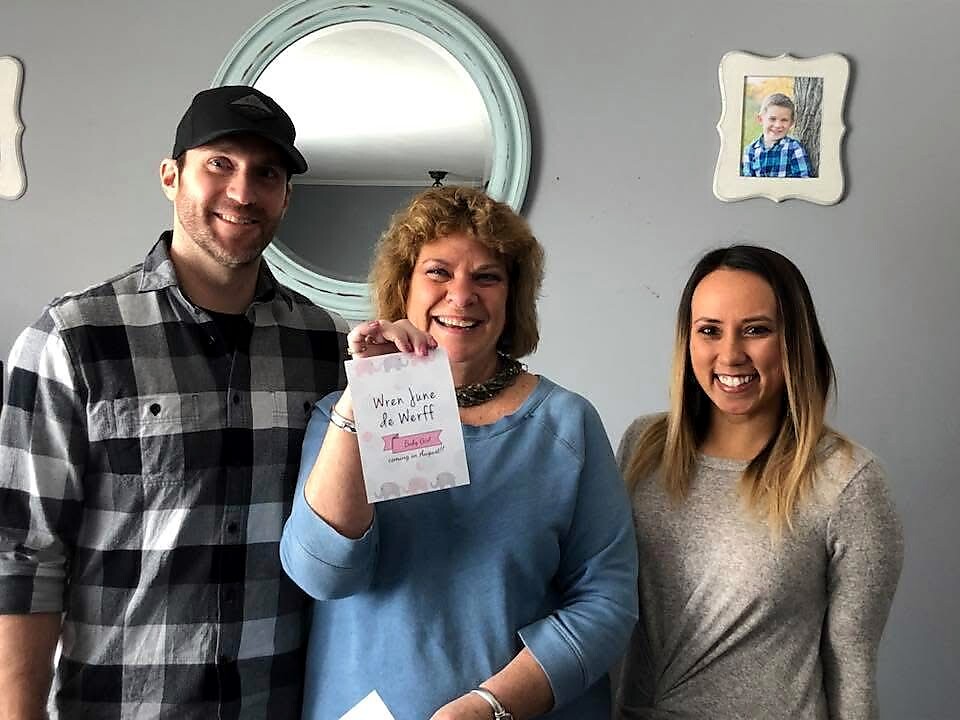 Three people smiling indoors; the center woman holds a card toward the camera.