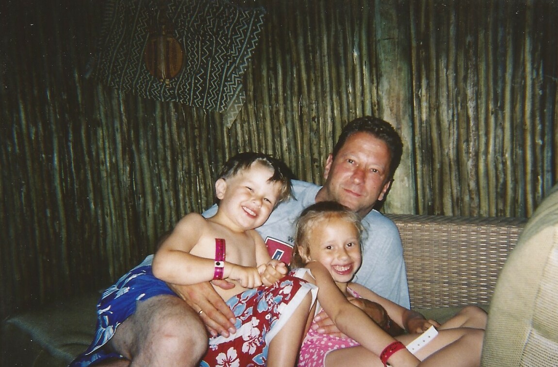 Father sits with two smiling children on a wicker sofa in a rustic, bamboo-walled room; kids wear colorful swimwear and bracelets.