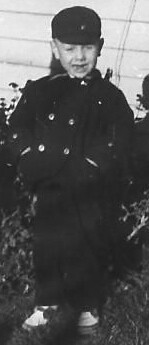 Black-and-white photo of a young child in a dark double-breasted coat and cap, standing outdoors among plants.
