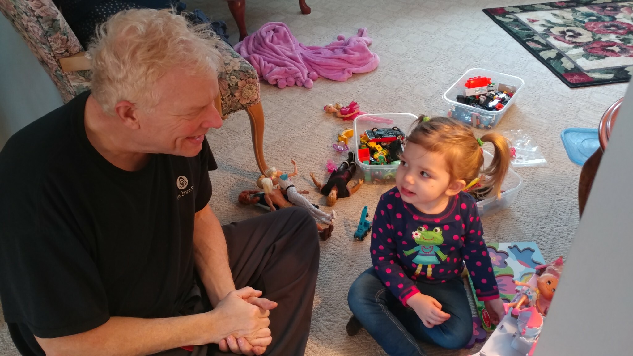 A man with gray hair and a young girl sit on a carpet, surrounded by toy figures and plastic bins of blocks, talking or playing.