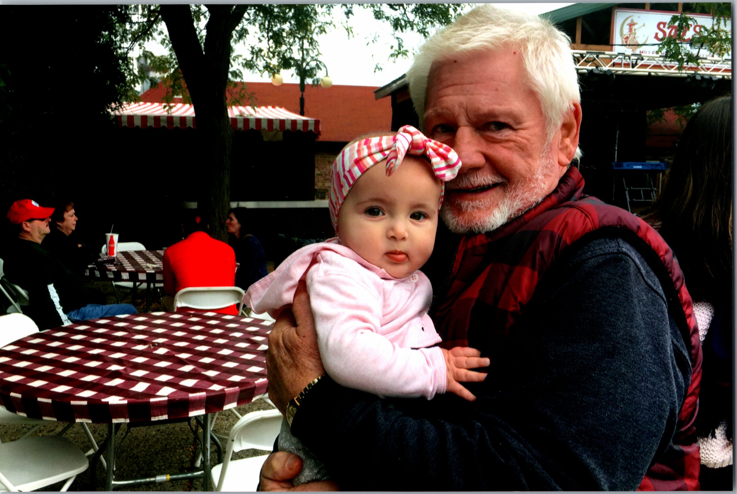 Older man with white hair smiles while holding a baby girl in pink at an outdoor cafe with red-and-white checkered tables.