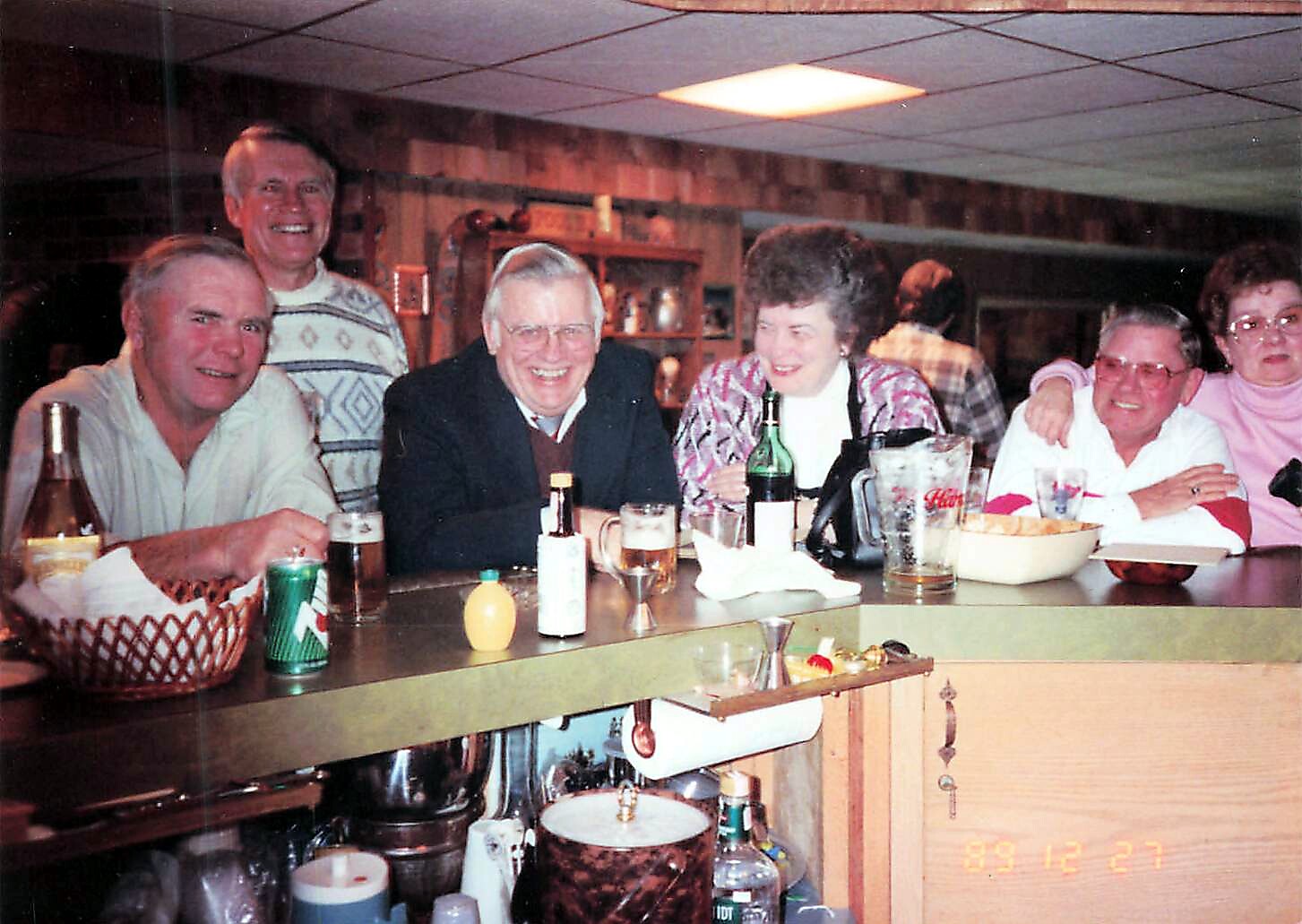 Six adults gathered at a bar smiling, with drinks and snacks on the counter in a cozy, retro setting.