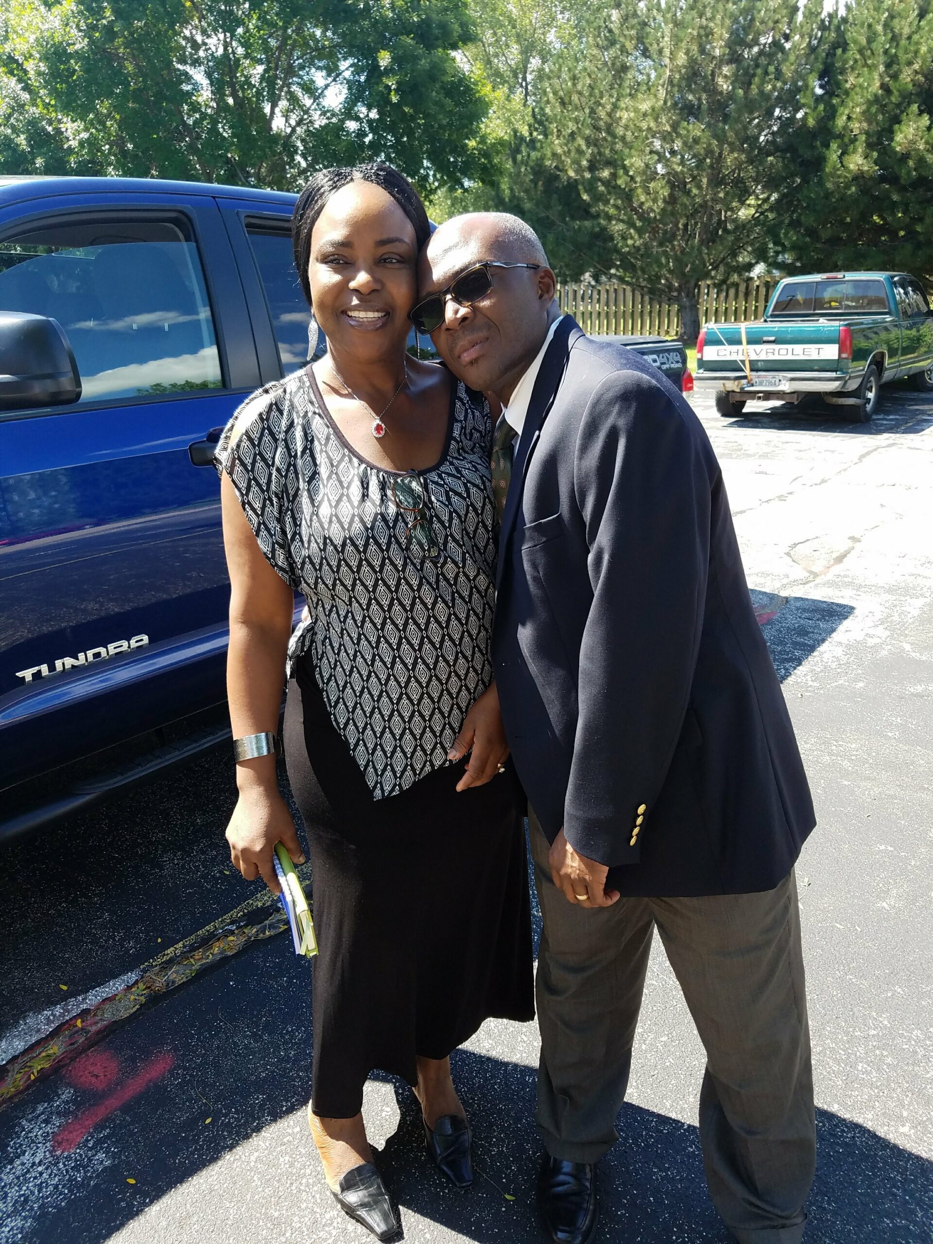 Smiling couple posing together outdoors beside a blue pickup truck in a parking lot; trees in the background.