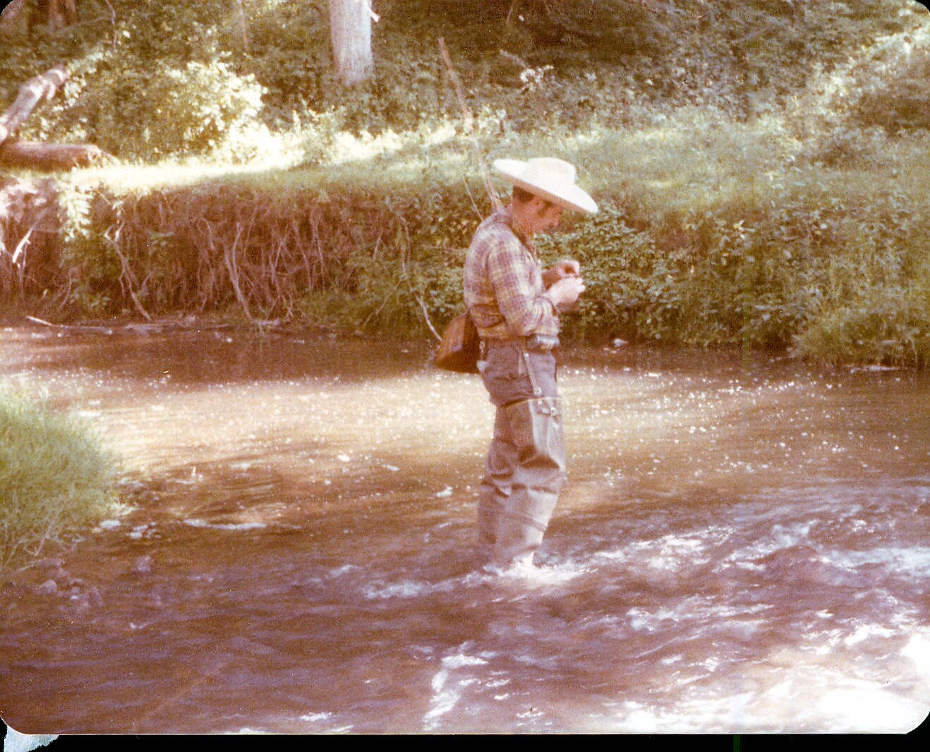 Man in a wide-brim hat and plaid shirt standing in a shallow river, fishing with a line.