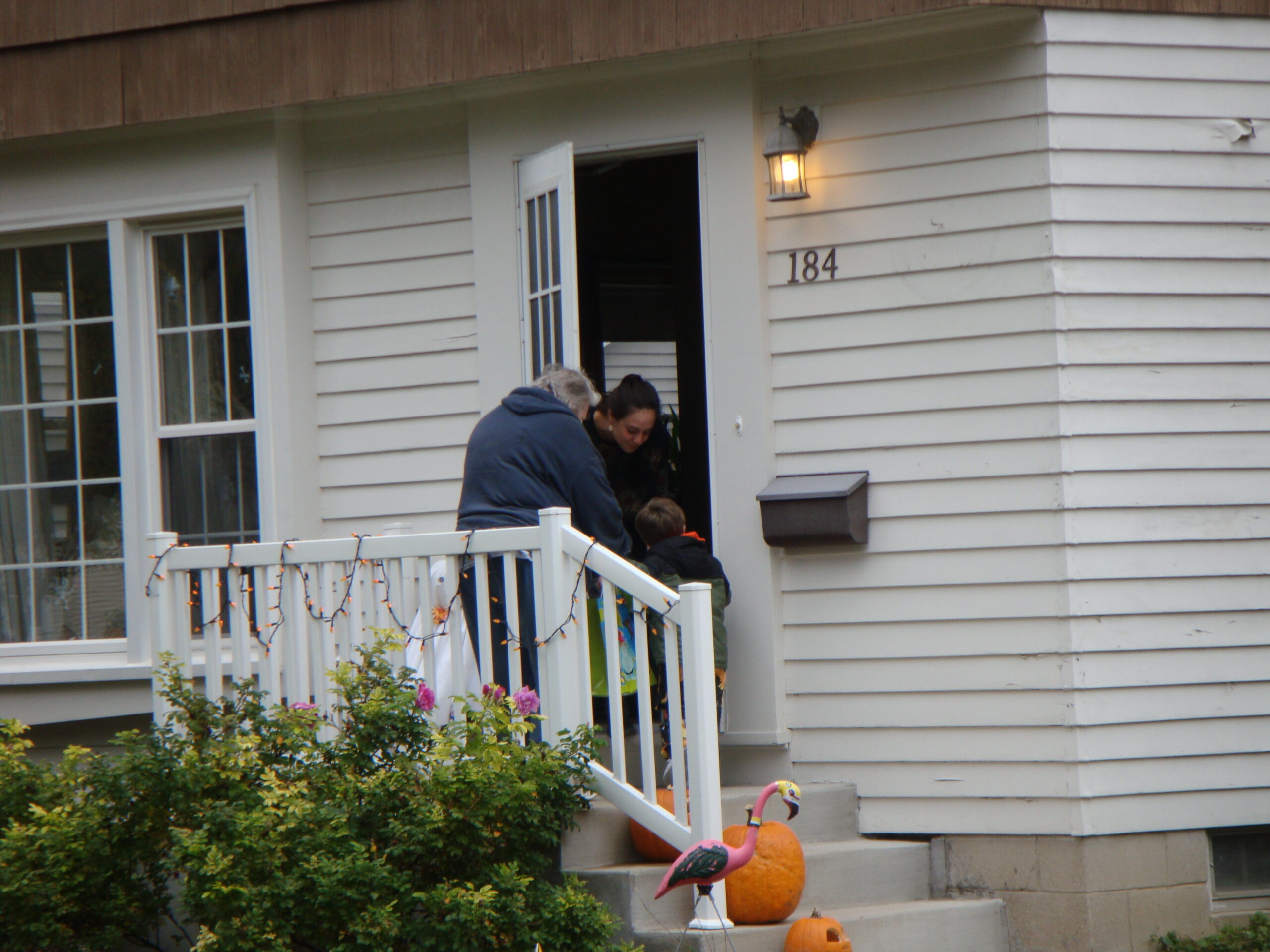 People stand on the front porch of a beige house with the door open, pumpkins on steps, and string lights along the railing (house number 184).