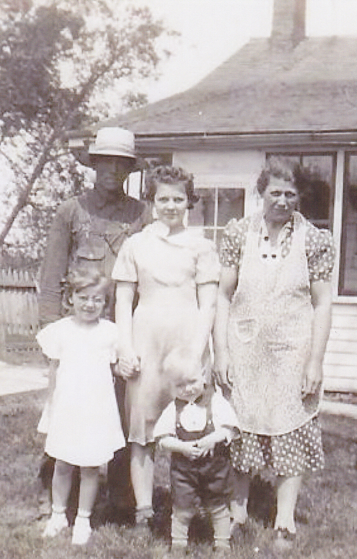 Family of five posing on a lawn in front of a house: man in a hat, two women in dresses, and two children