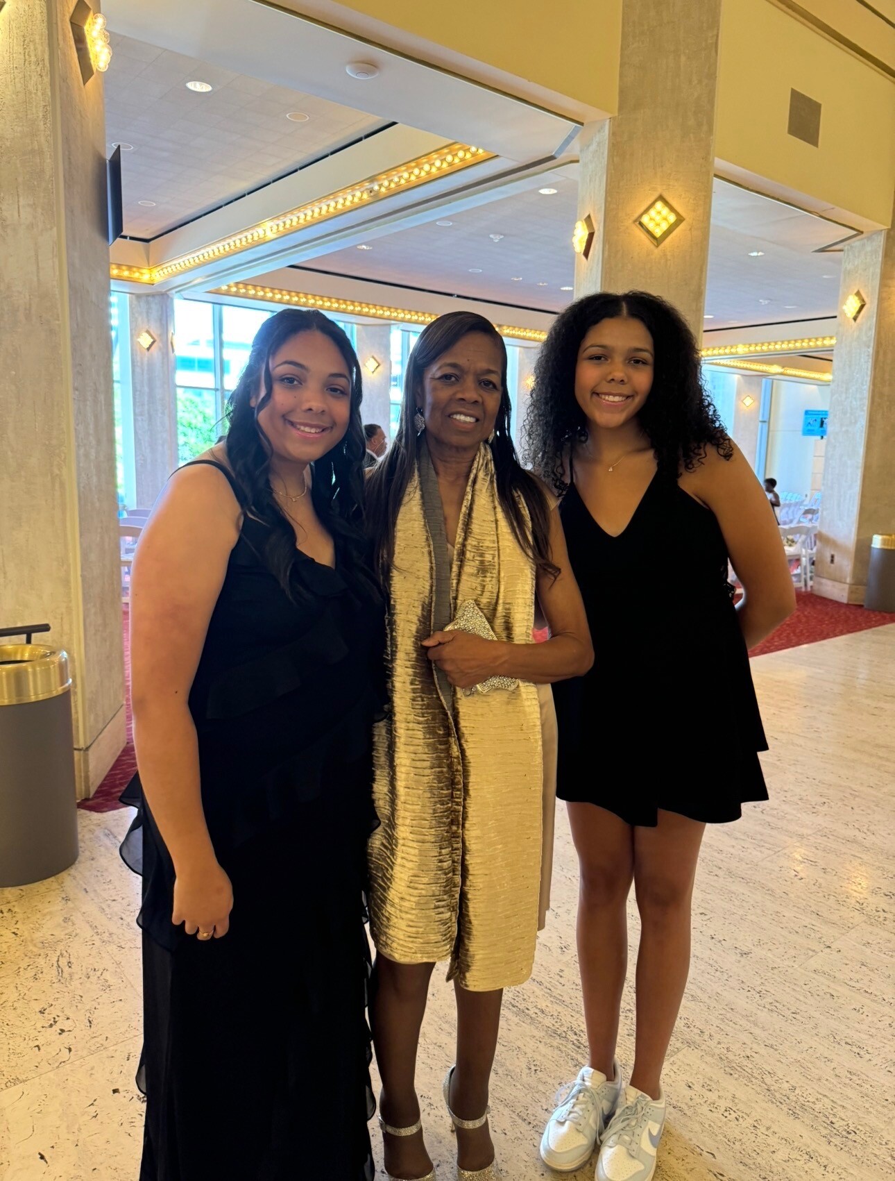 Three women in black dresses pose together in a lobby with gold-toned lighting and marble columns.