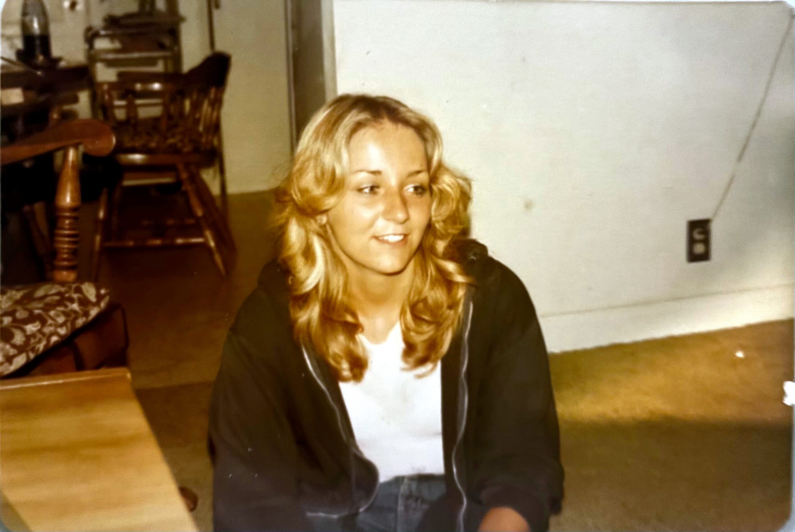 Young woman with blonde hair sitting on the floor indoors, smiling at the camera in a living room setting.