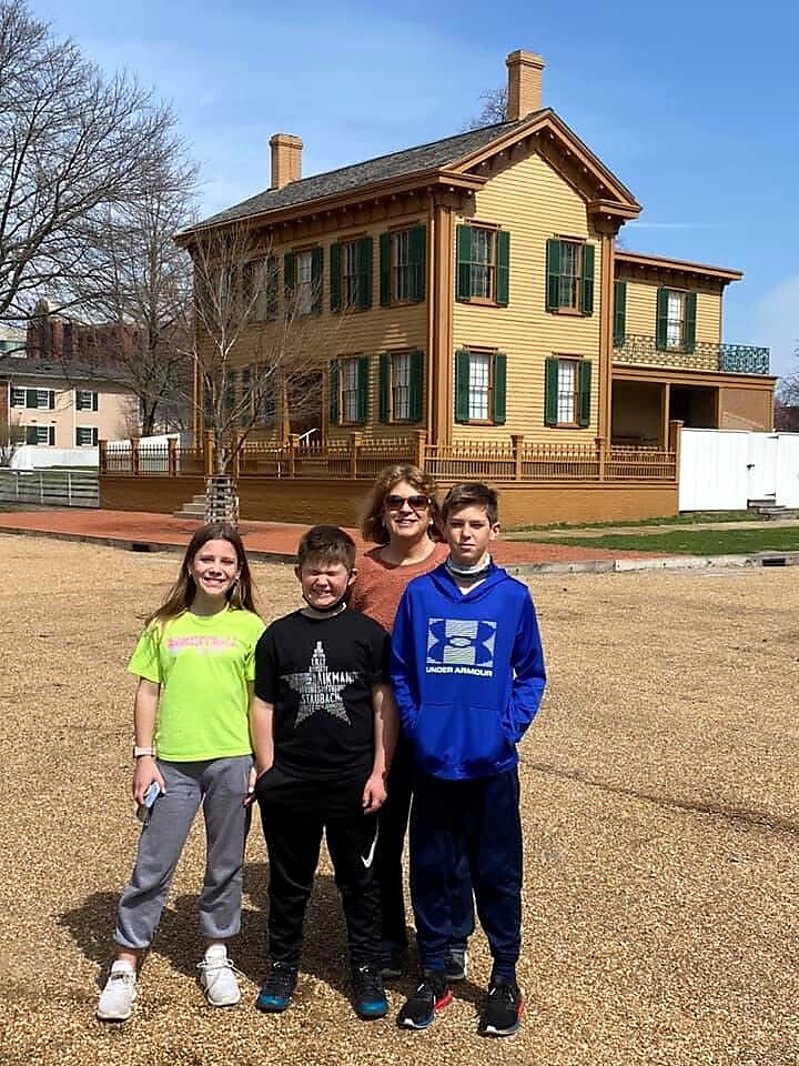 Four kids and an adult woman pose on gravel in front of a large yellow house with green shutters on a sunny day.