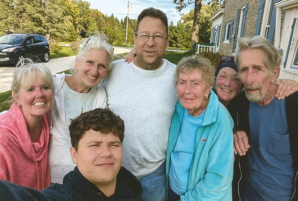 Seven people pose for a selfie outdoors on a sunny residential street, all smiling at the camera.