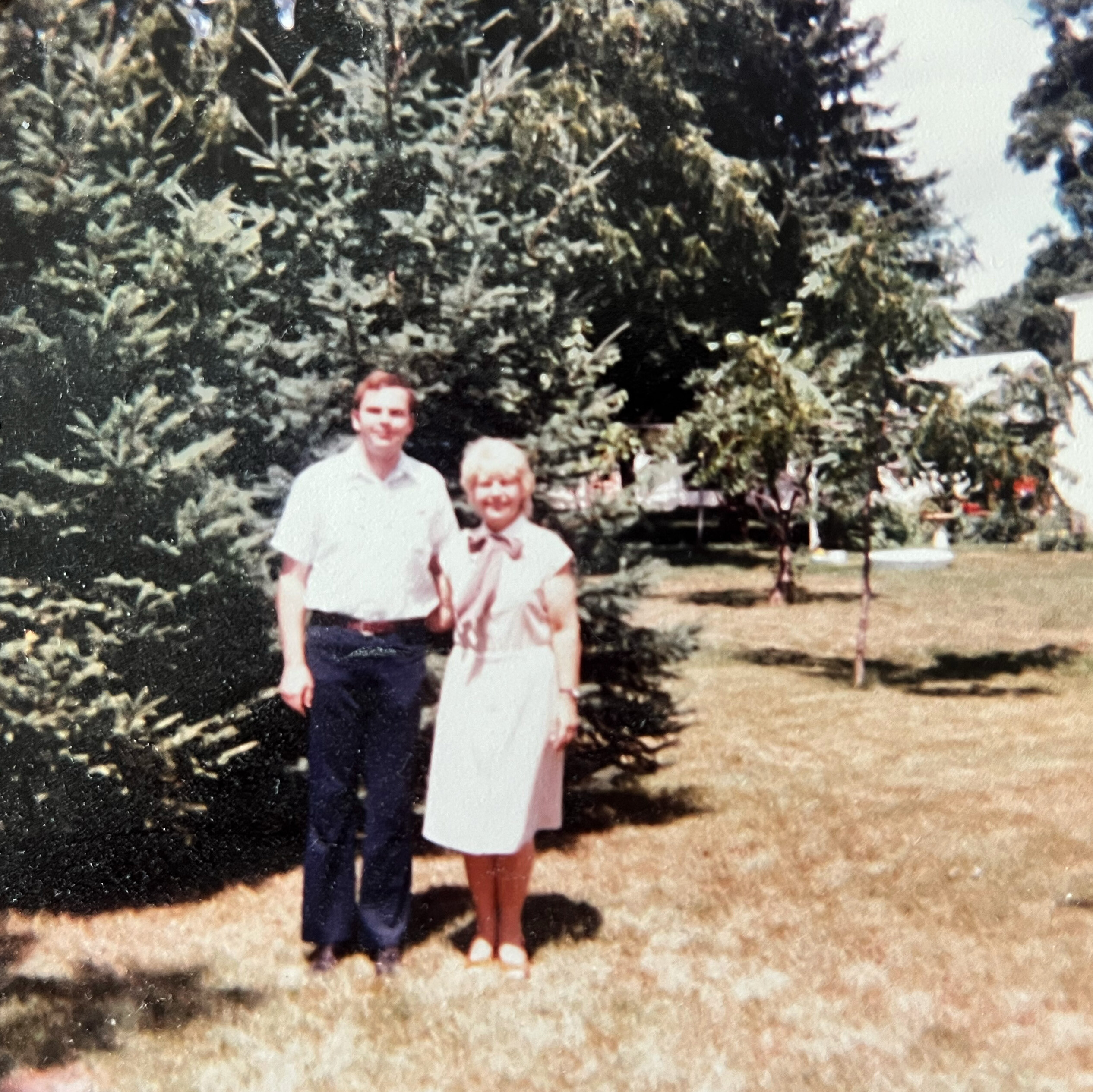 Two people stand outdoors in front of a large shrub, posing for a family photo on a sunny day.