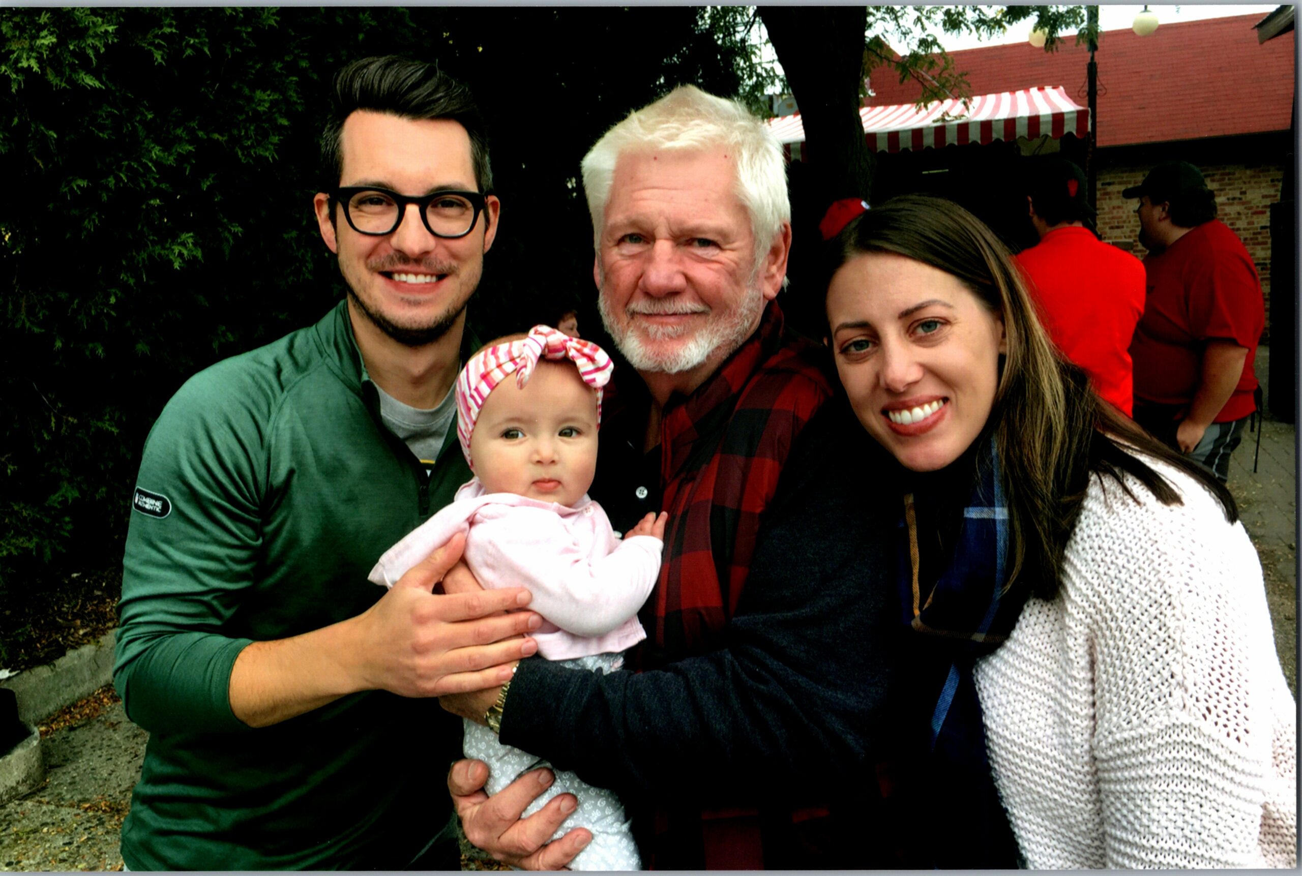 Smiling family of four posing outdoors with a baby girl, man with glasses, an older man, and a woman; red-and-white striped tent in the background.