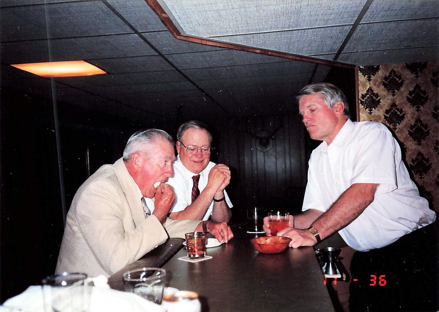 Three older men sit at a bar chatting and eating, with drinks on the counter; the man on the left bites into a snack, the middle man smiles, and the right man in a white shirt leans on the bar.