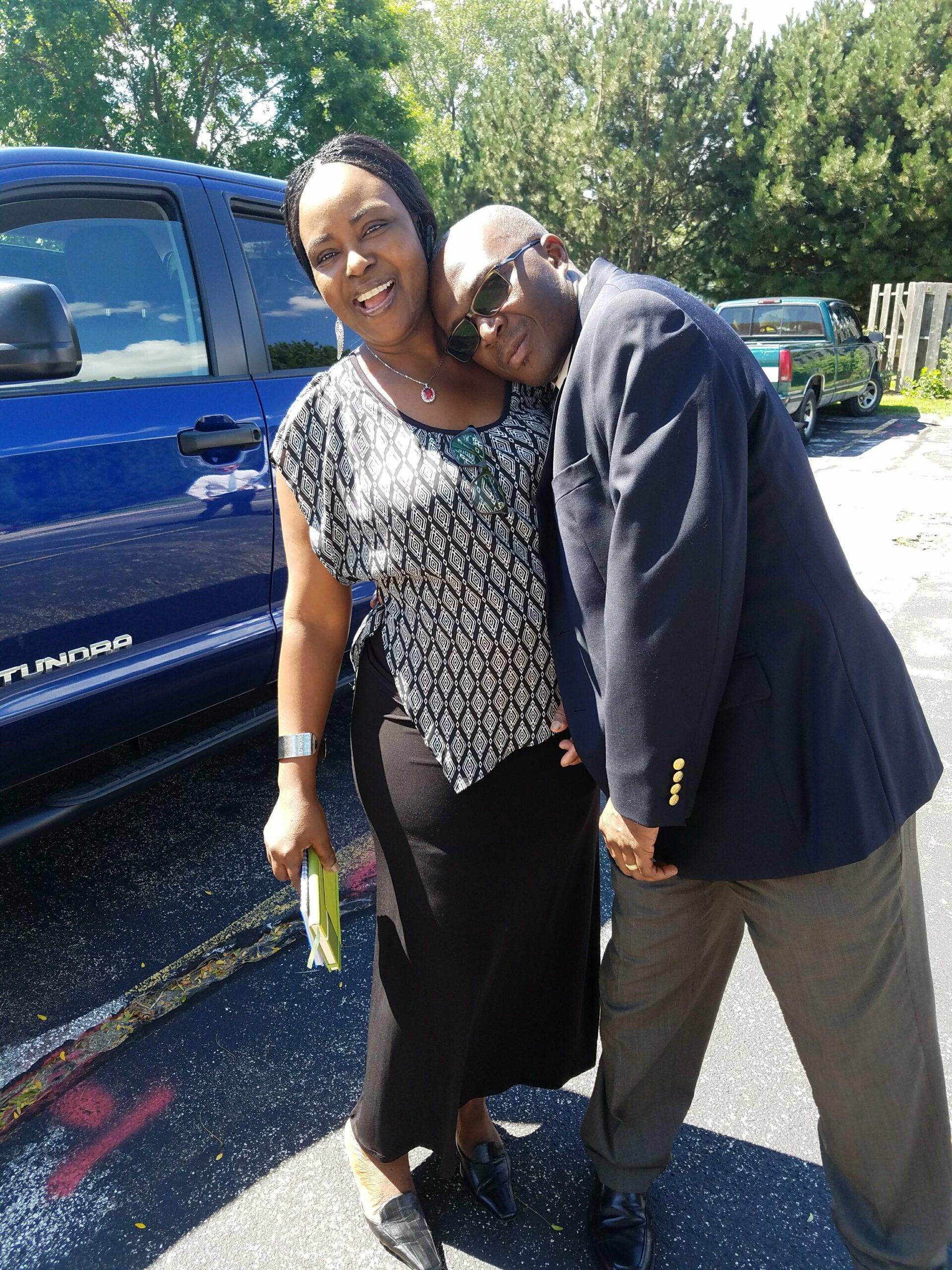 Couple posing together by a blue SUV, the man resting his head on the woman's shoulder while wearing sunglasses and a navy blazer.