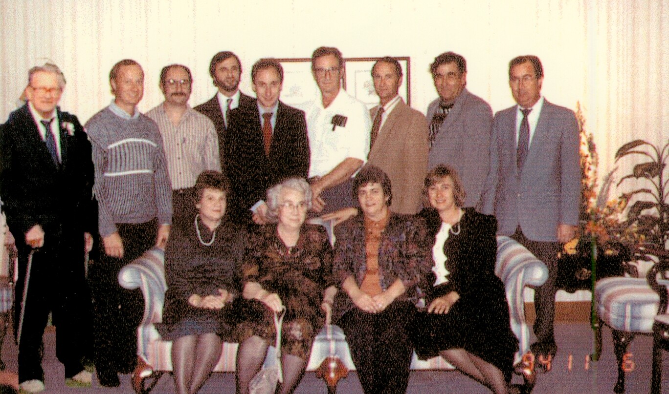 Twelve adults posing for a group photo in a living room, some seated on a sofa and others standing behind them.
