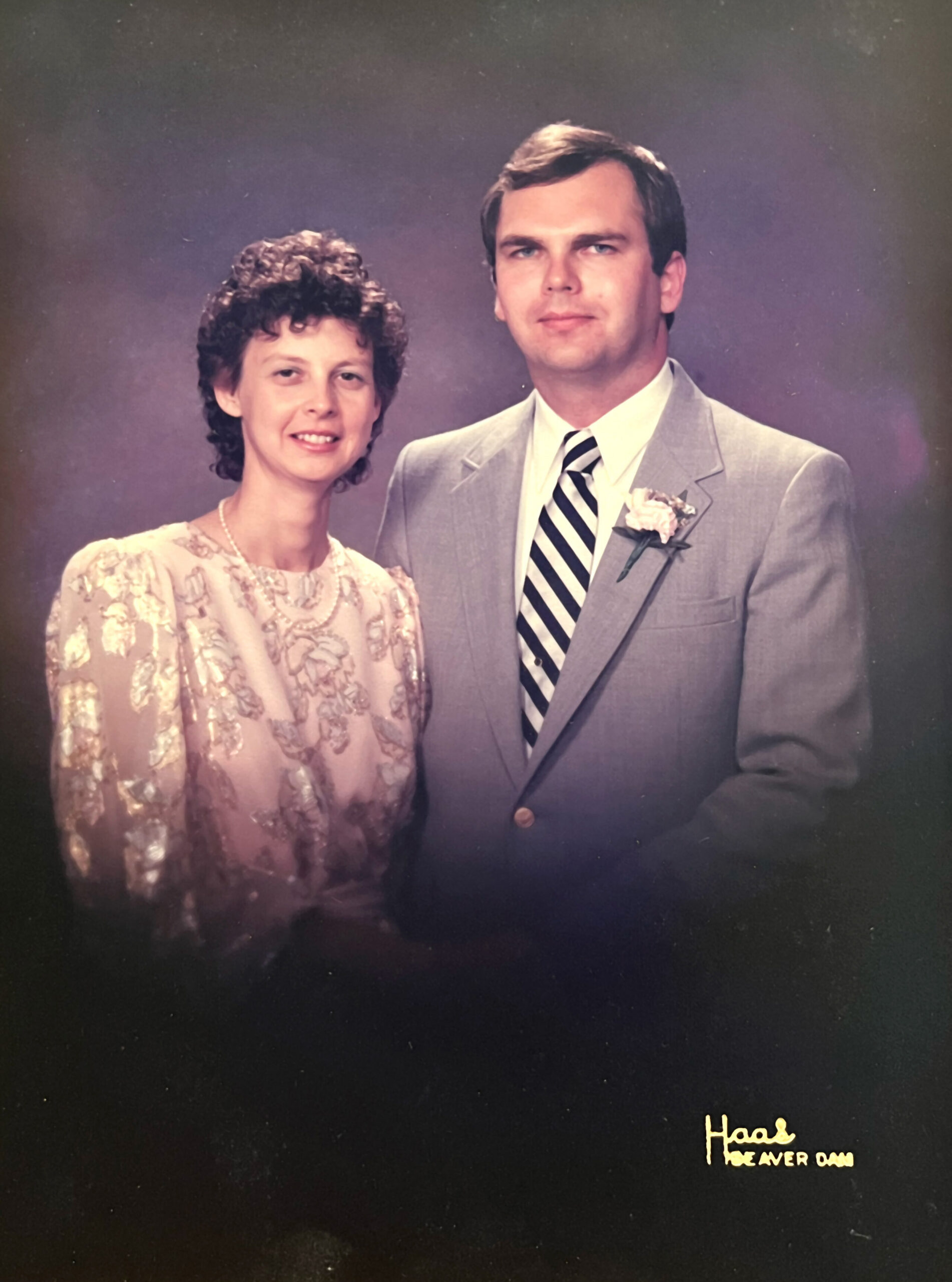 Portrait of a couple in formal attire: woman in a lace dress and pearl necklace, man in a light gray suit with a striped tie and boutonniere, against a studio background.