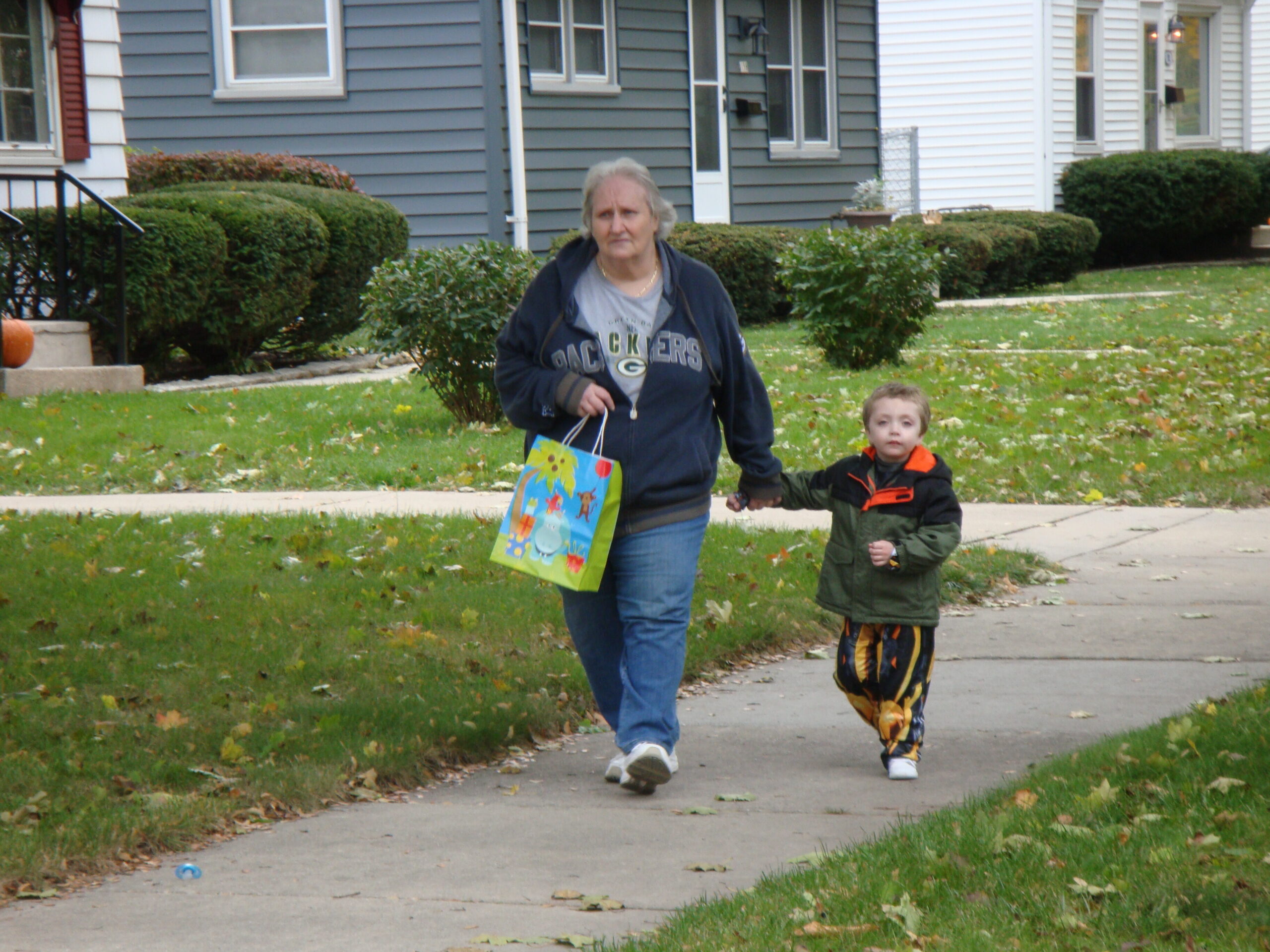 An older woman and a young child walk hand in hand along a suburban sidewalk on a fall day, carrying a colorful gift bag.