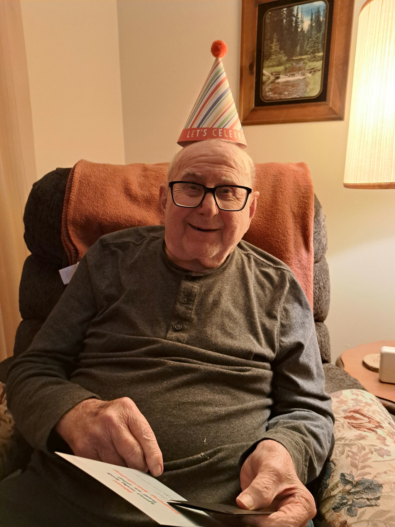 Elderly man with glasses wearing a colorful party hat that reads 'Let's Celebrate', sitting in a chair and looking at a card he’s holding.