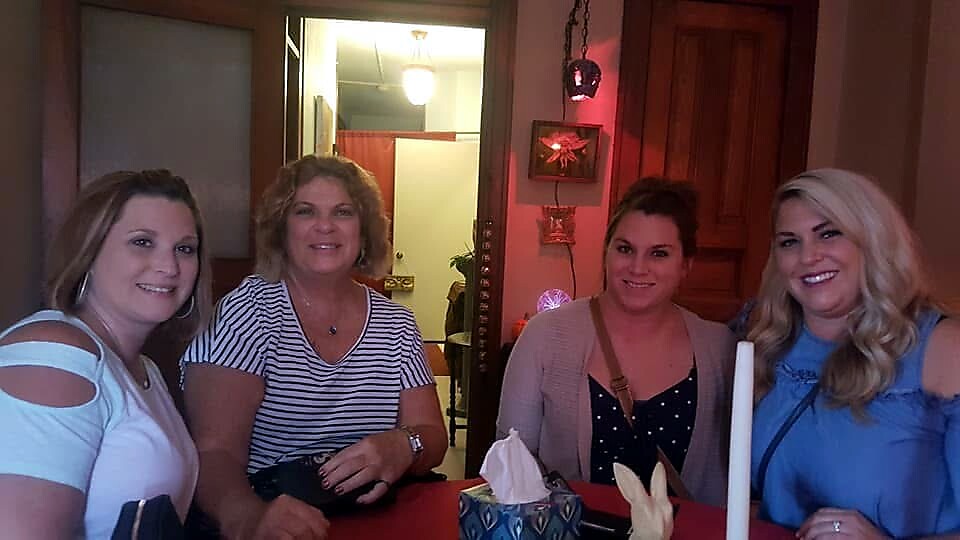 Four women smile at a table in a cozy room, posing for a photo together with a tissue box and candle on the table behind them.