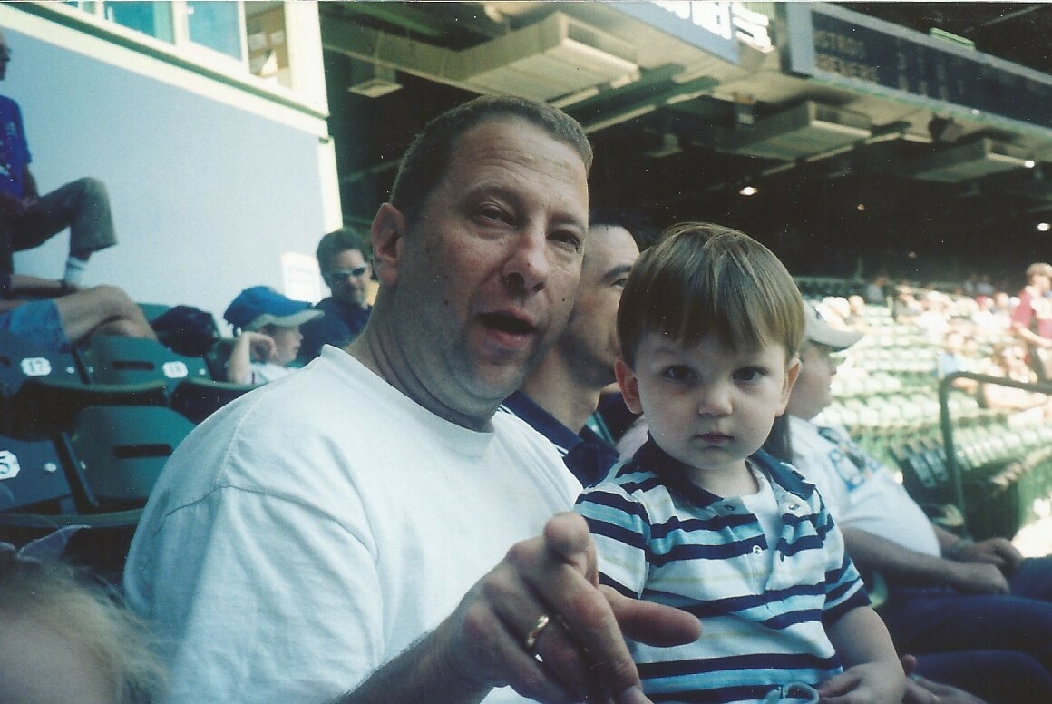 Man in a white shirt and a toddler in a striped polo sit in stadium seats; the man points at the camera.