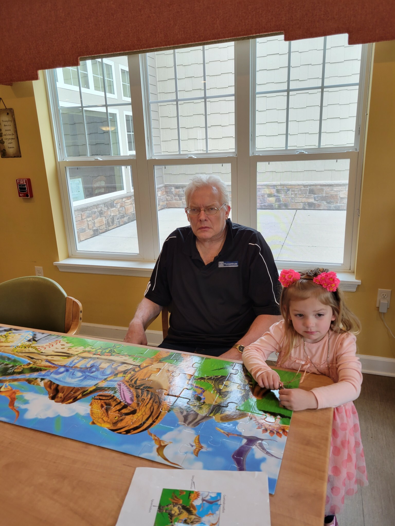 Older man with white hair and glasses seated at a table assembling a bright dinosaur puzzle, with a young girl nearby.