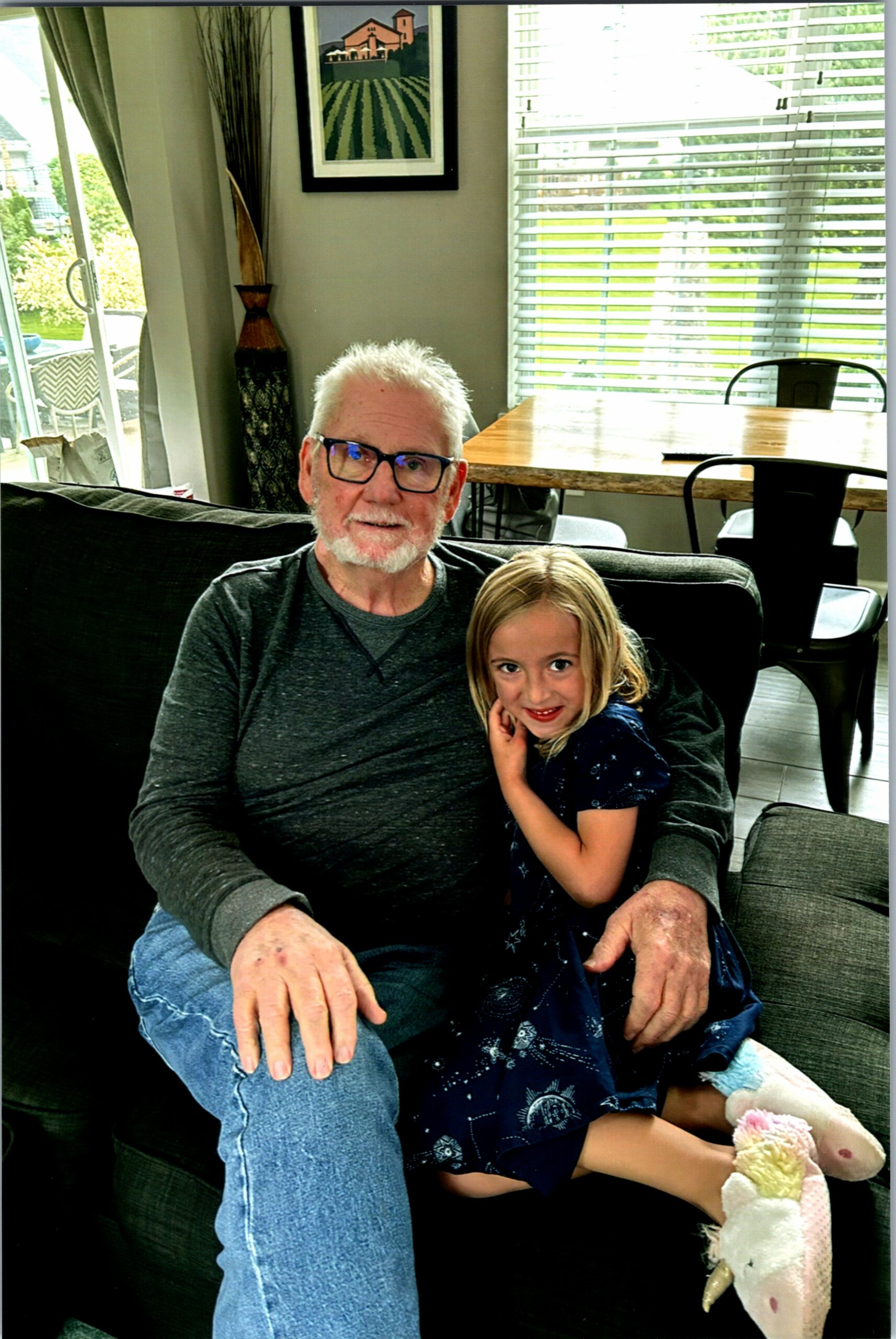 Older man with white hair and glasses sits on a sofa, smiling as a young girl hugs him.