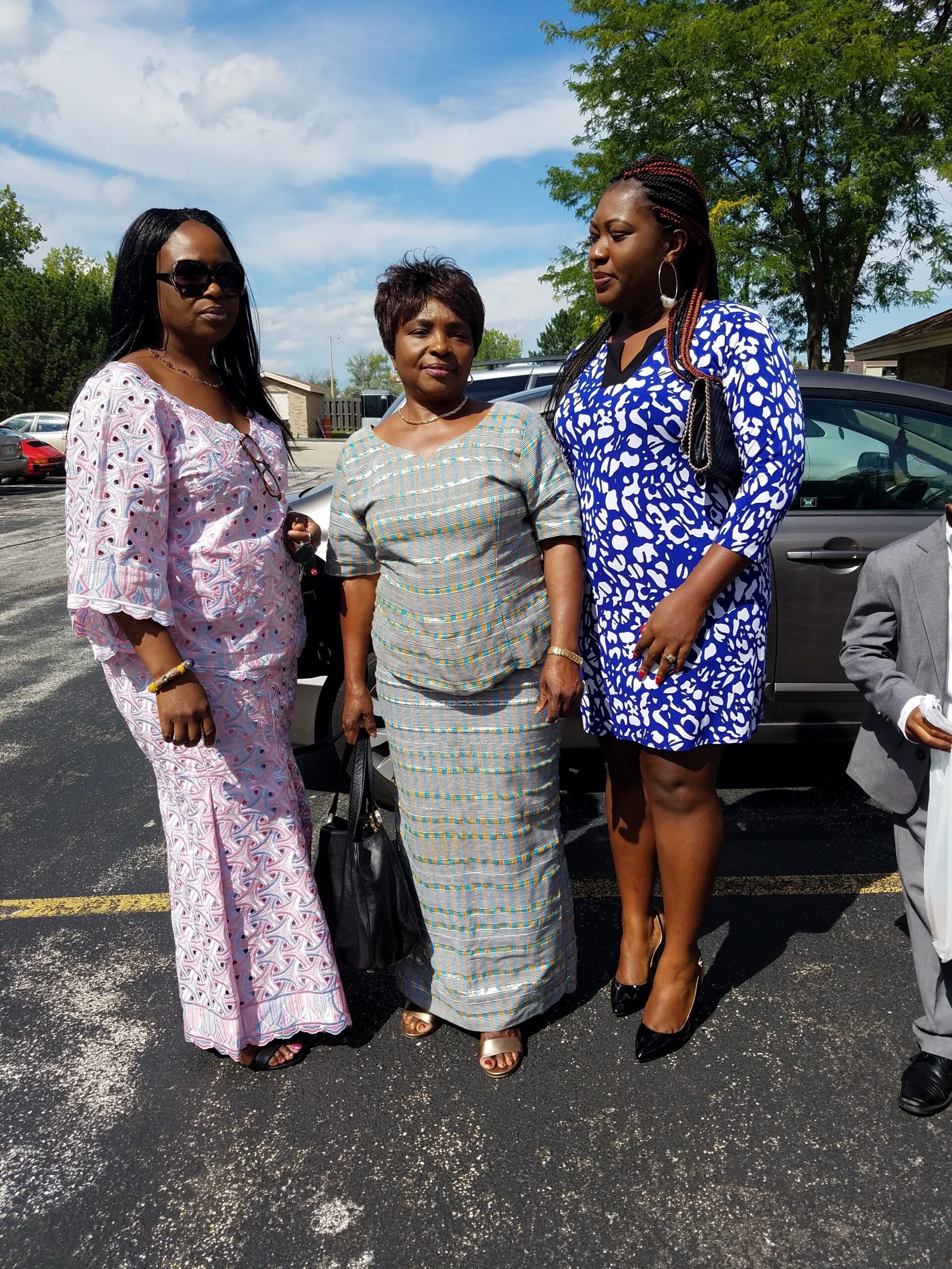 Three women posing together in a parking lot on a sunny day, wearing colorful dresses and accessories (pink pattern, striped, and blue/white print).