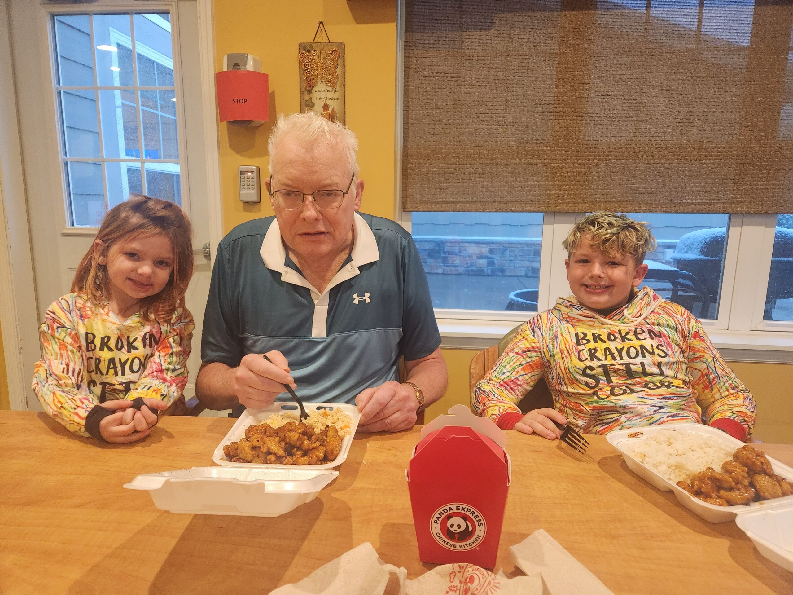 Three people — an older man and two children — sit at a table eating takeout, with a Panda Express cup in the foreground and food trays visible; the kids wear colorful tie-dye shirts that read 'Broken Crayons Still Great' and a snowy window is in the background.