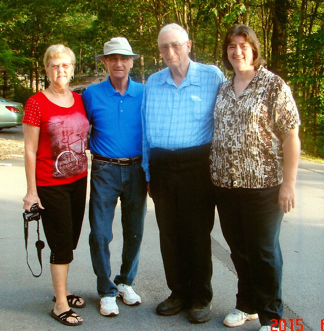 Four adults pose together outdoors on a sunny street, smiling, with trees in the background; woman on left holds a camera.