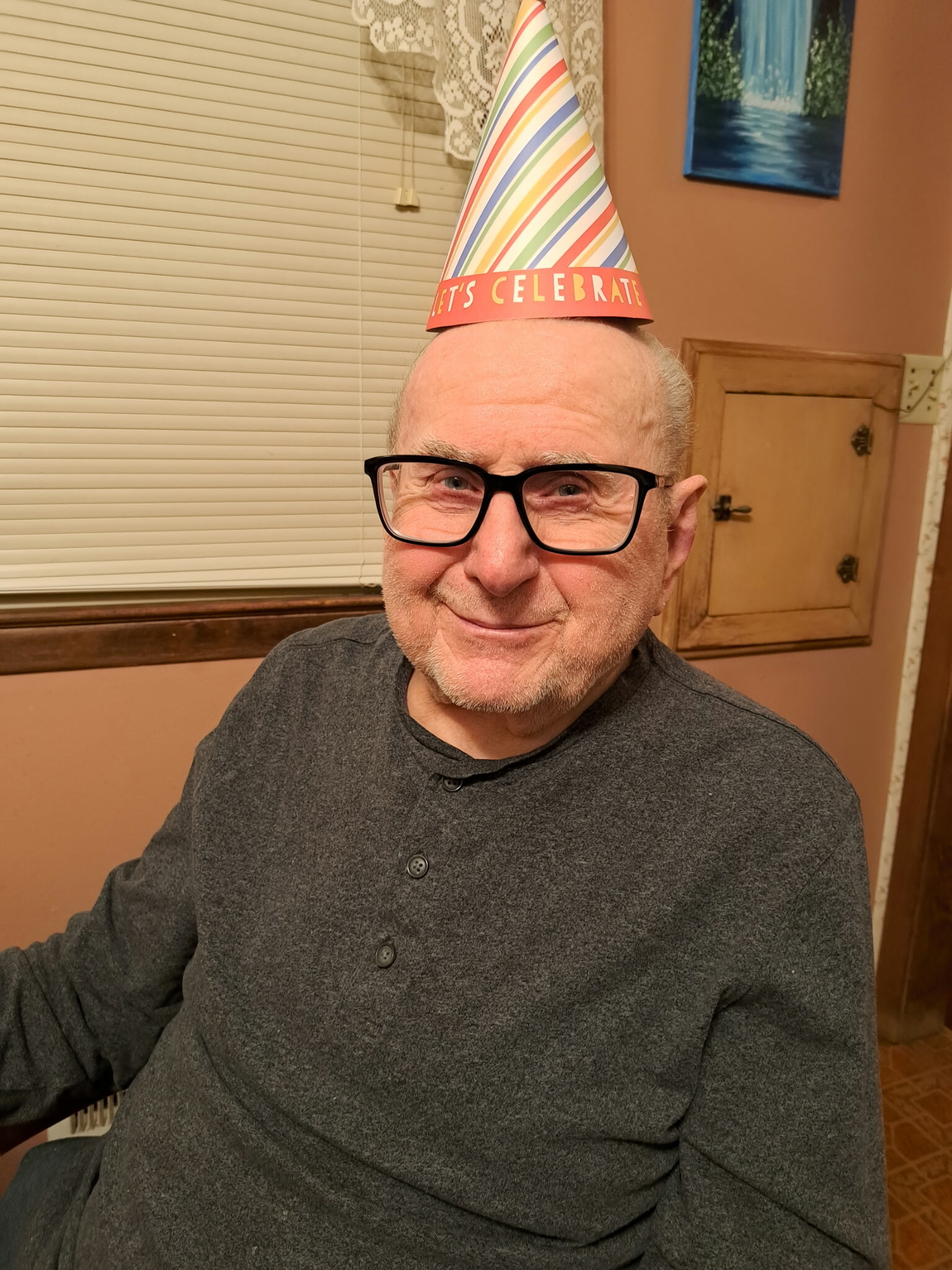 Older man with glasses wearing a colorful birthday party hat that says 'Let's Celebrate' indoors, smiling at the camera.