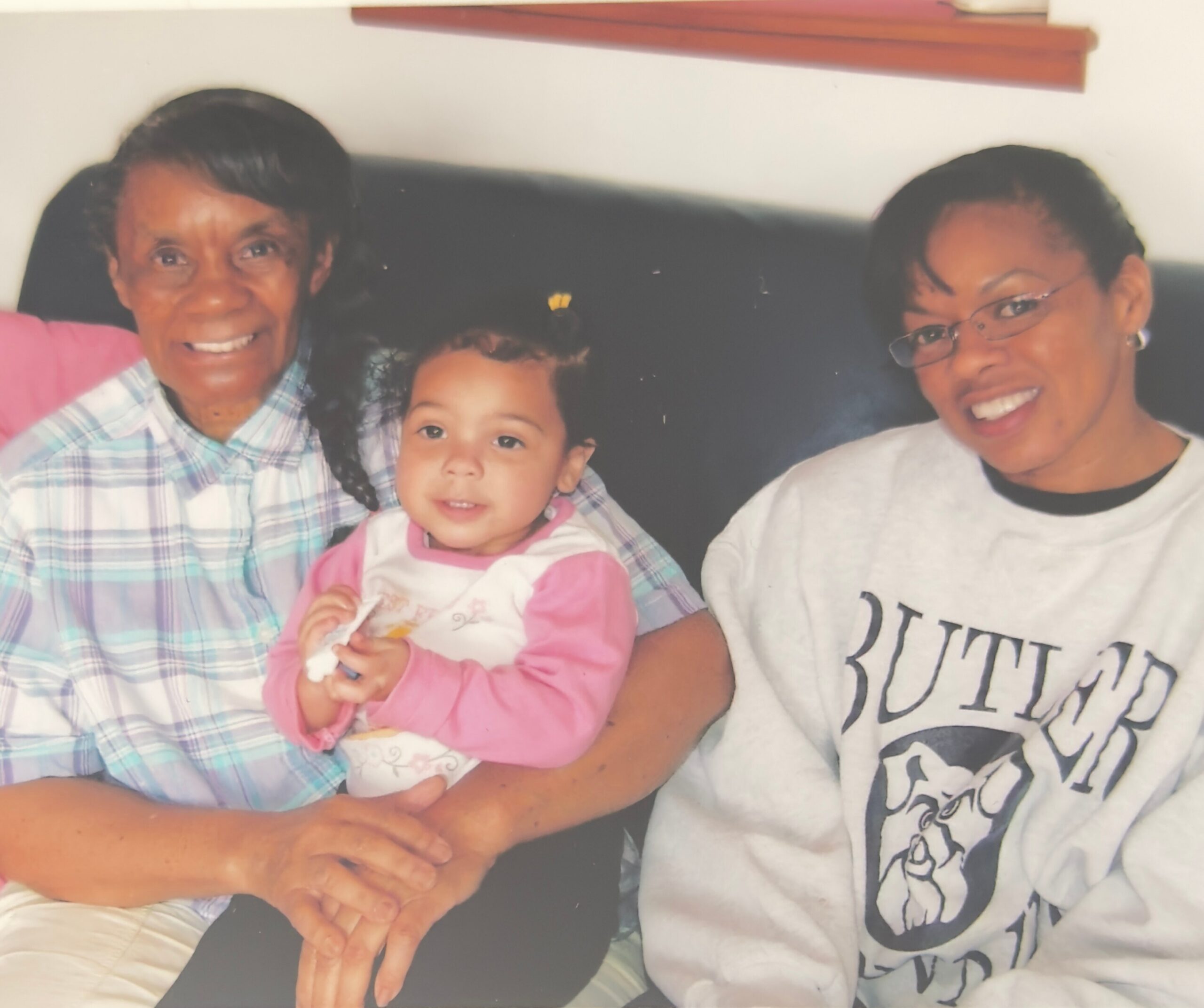 Three generations of a family sitting on a couch: an elderly woman on the left, a baby in pink in the middle, and a smiling woman with glasses on the right.