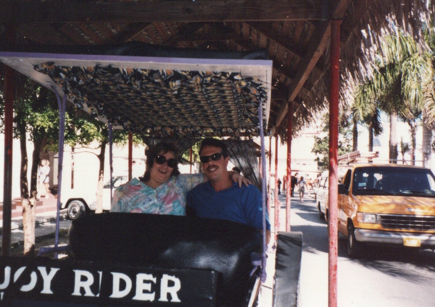 Two people wearing sunglasses ride in a open trolley on a sunny street lined with palm trees; a partial sign reads 'JOY RIDER' at the front.
