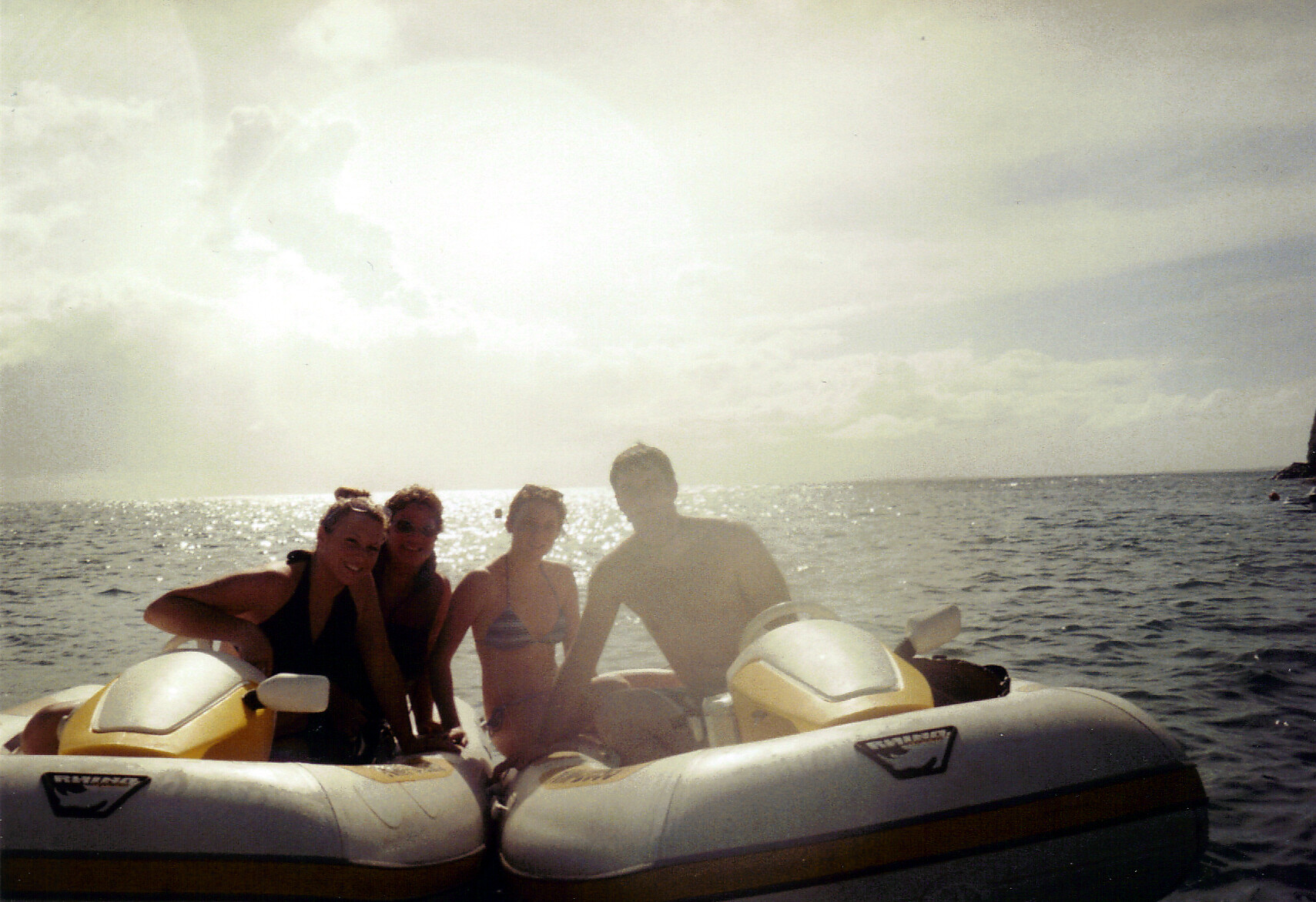 Four friends on a yellow-and-white inflatable raft in the sea, wearing swimsuits, smiling at the camera as the sun glints on the water.